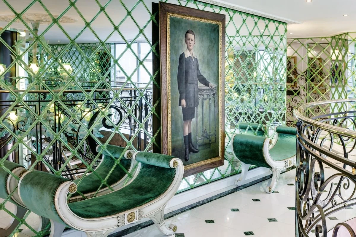Interior of a luxurious space featuring a large portrait of a young woman, ornate green velvet benches, decorative green lattice partitions, and elegant wrought iron railings.