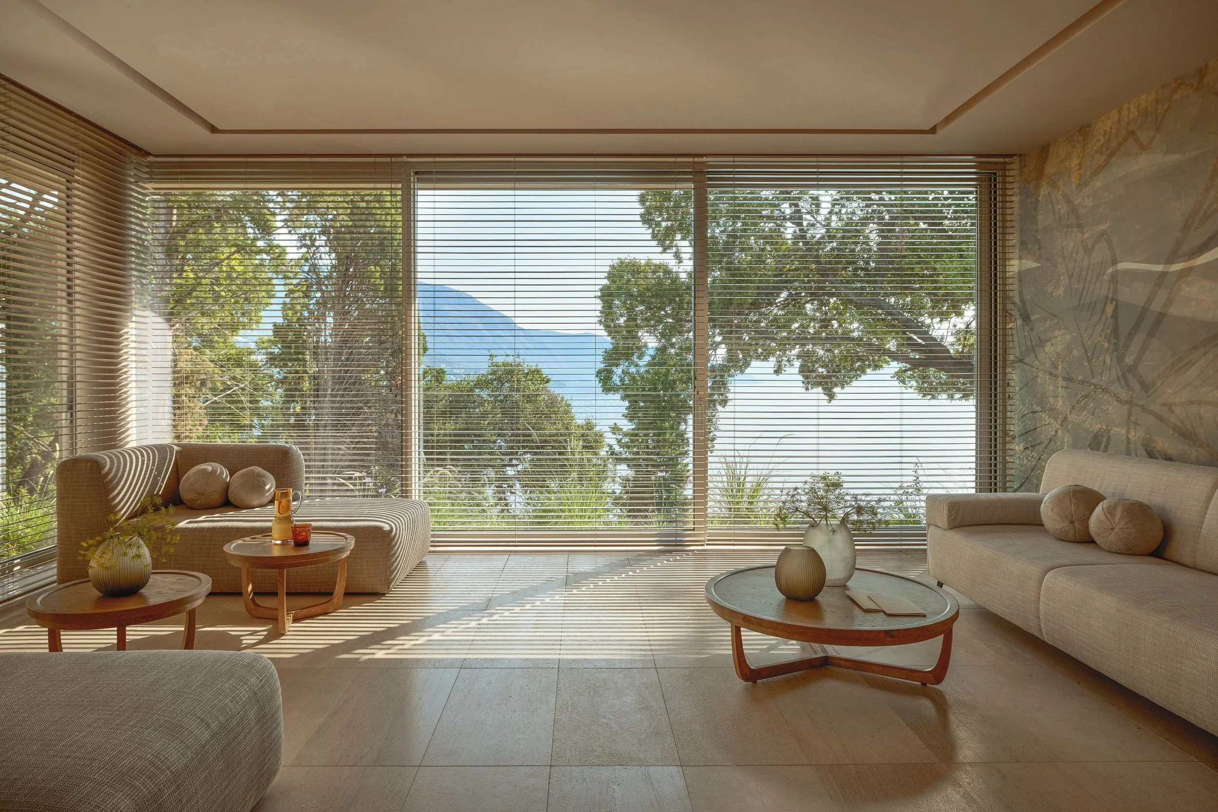 Living room with large floor-to-ceiling windows, sunlight casting shadows, modern beige sofa with round cushions, round coffee tables with decorative vases, and a view of trees and mountains outside.