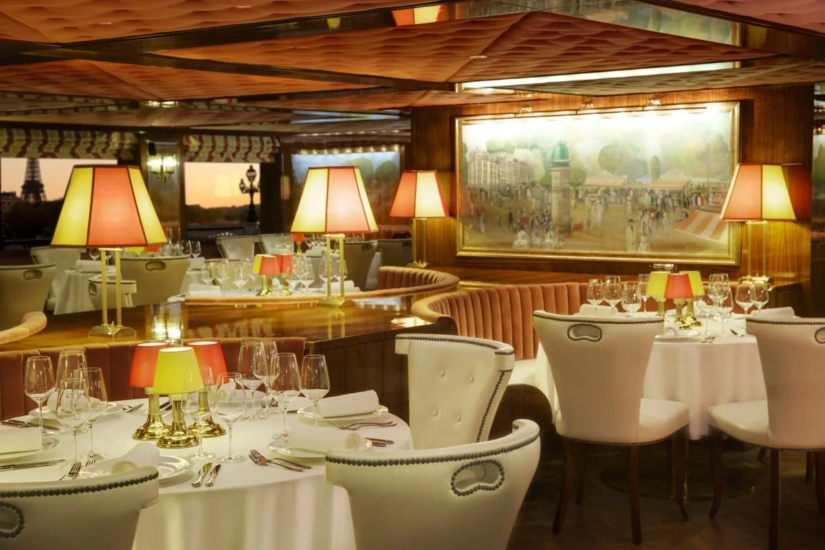Elegant restaurant dining area with round tables set with white tablecloths, wine glasses, and napkins, illuminated by yellow and red lamps, with plush white chairs, a large mirror, and window views of a bridge and cityscape at sunset.