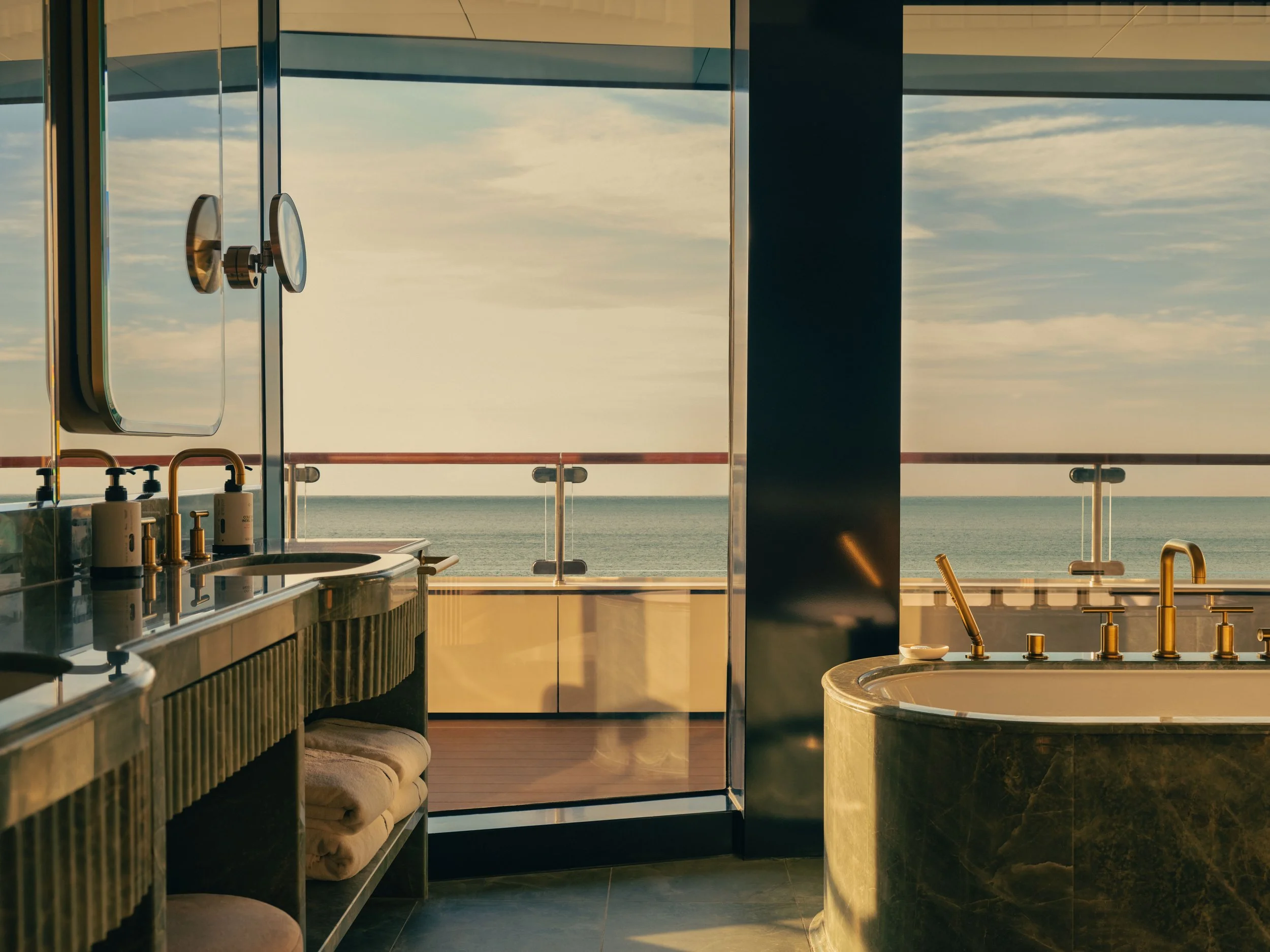 A luxurious bathroom with a view of the ocean, featuring a double sink vanity with soap dispensers, a glass shower door, a round bathtub, and large windows showing a cloudy sky and sea.