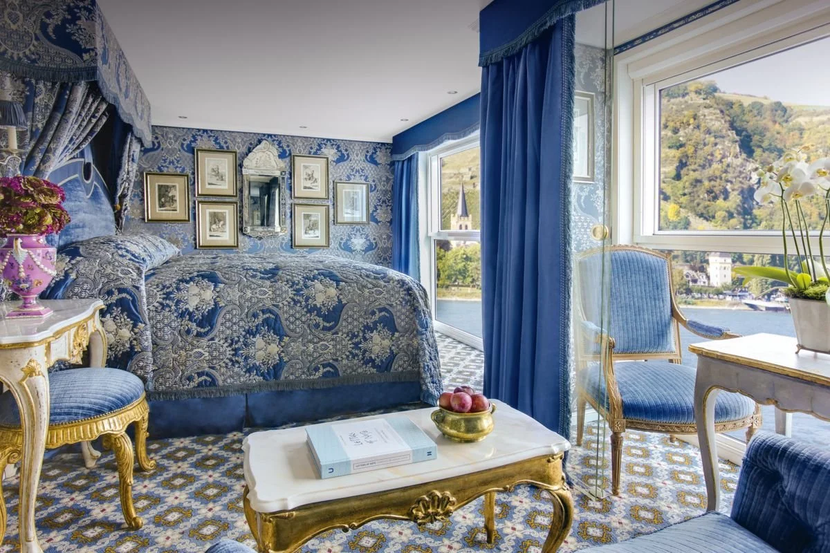 A luxurious bedroom with blue and white decor, including patterned curtains and bedding, gold-trimmed furniture, a mirror, framed artwork, and a large window overlooking a scenic view of water, hills, and a church steeple.