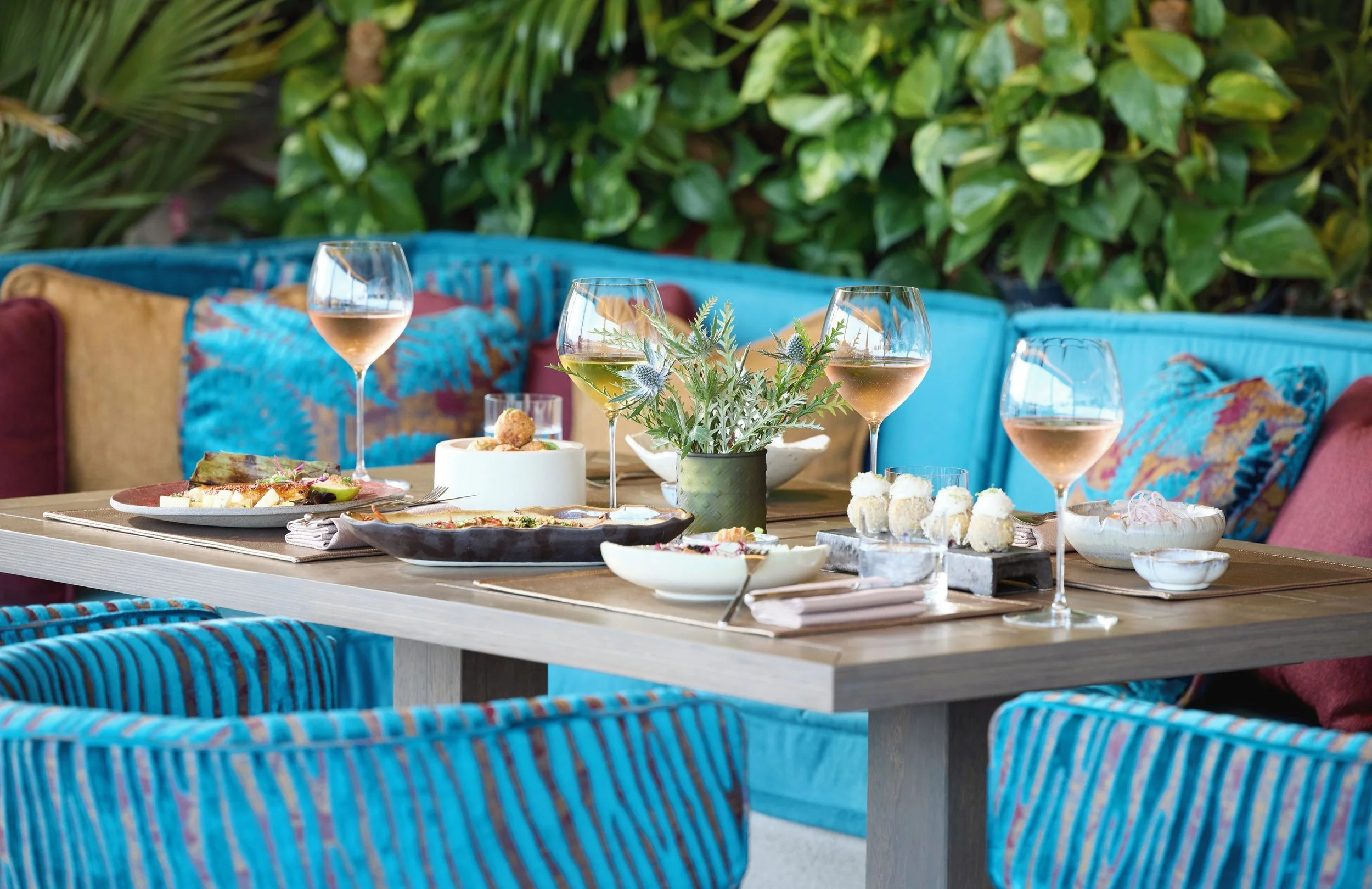 A dining table set outdoors with various foods, wine glasses, and a potted plant, surrounded by colorful woven chairs and lush green foliage.