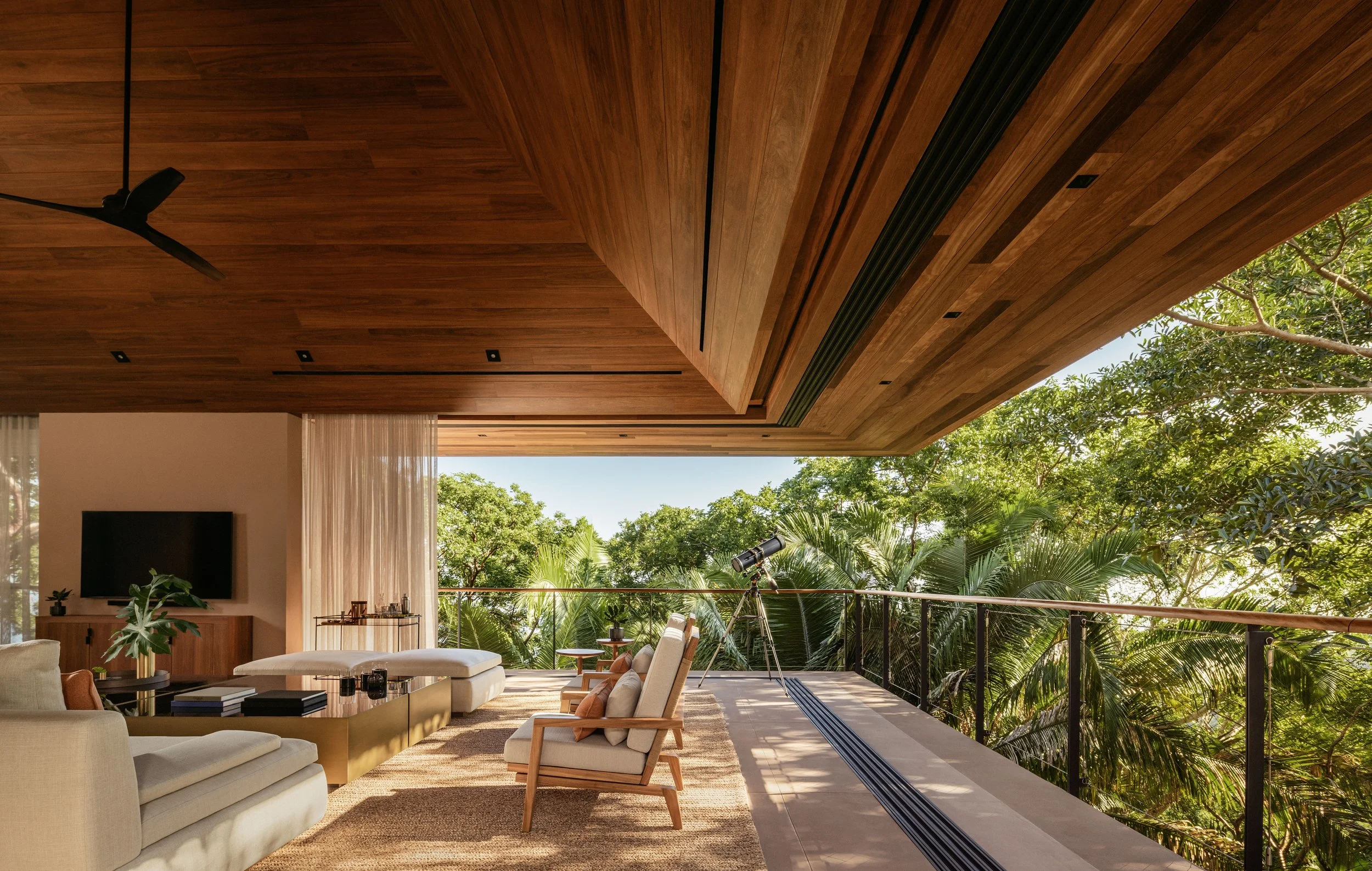 Modern indoor living space with large open windows, wooden ceiling, ceiling fan, and lush greenery outside.