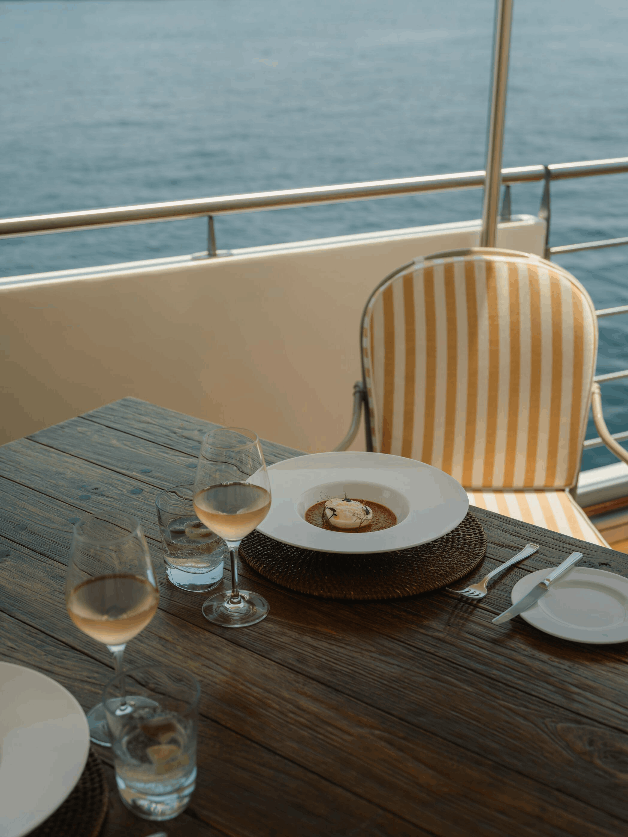 A dining table set with glasses of wine, a dessert, and silverware on a rustic wooden table, overlooking the ocean with a balcony railing in the background.