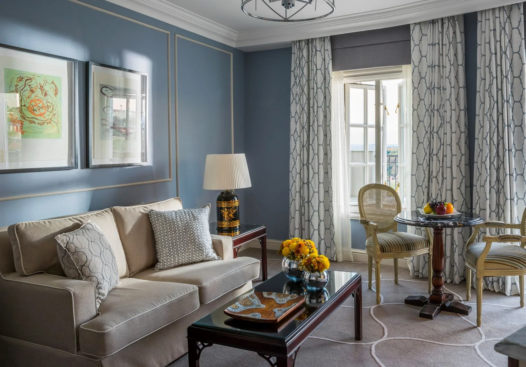 A charming living room with light blue walls, a beige sofa with patterned cushions, a wooden side table with a decorative lamp, and framed artwork on the wall. There are large windows with white and patterned curtains, and a small round table with a fruit bowl and chairs nearby. A coffee table with a floral arrangement is also visible.