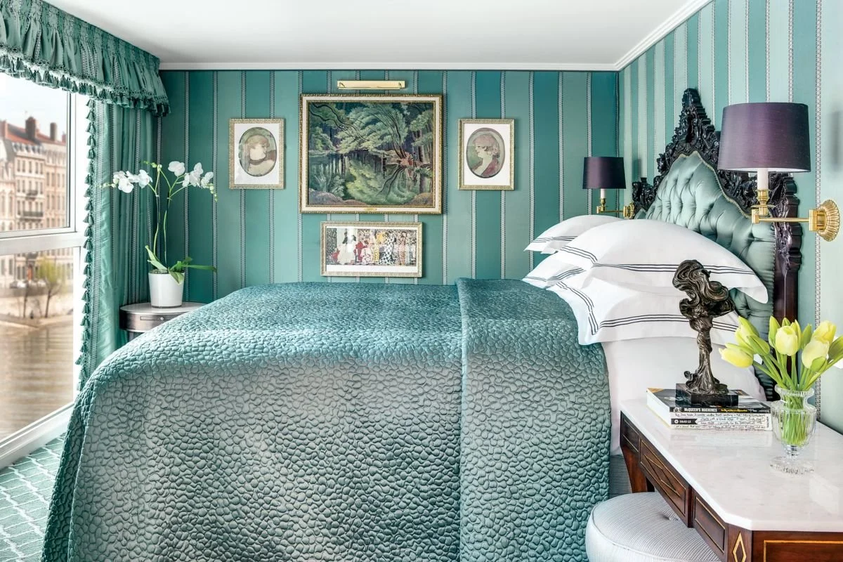 A bedroom with green and blue vertical striped wallpaper, a bed with a green tufted headboard, white sheets, and a textured teal bedspread. There are purple lamps on each side of the bed, a nightstand with a white marble top, a white orchid plant, and a window with green curtains showing buildings and a waterway outside. Framed artwork decorates the wall above the bed.