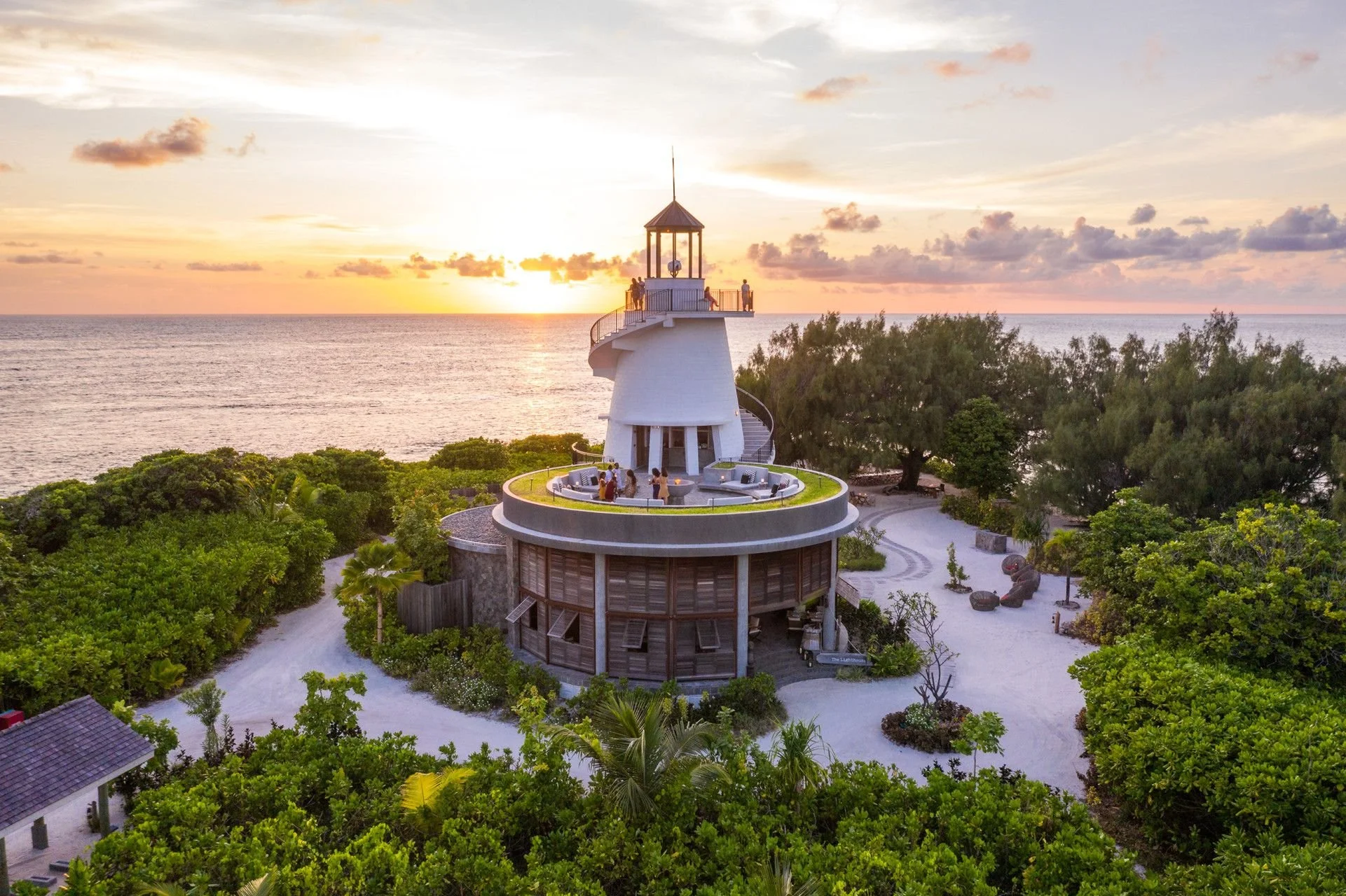 A white lighthouse with a small observation deck at the top, overlooking the ocean during sunset, surrounded by lush green trees and a sandy beach.