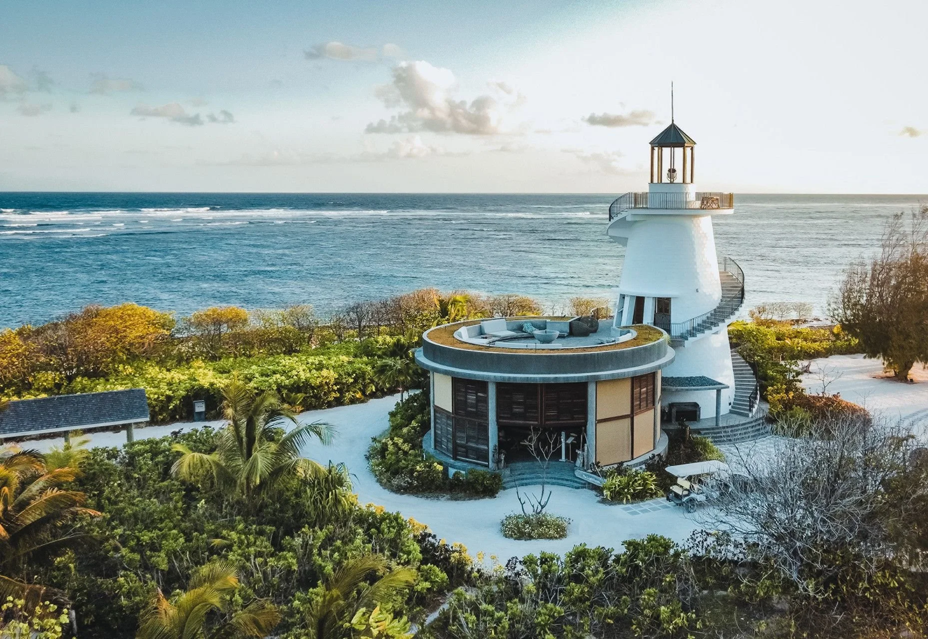 A white lighthouse with a spiral staircase, balcony, and top lantern located on a coastal landscape with ocean, cloudy sky, greenery, and trees.