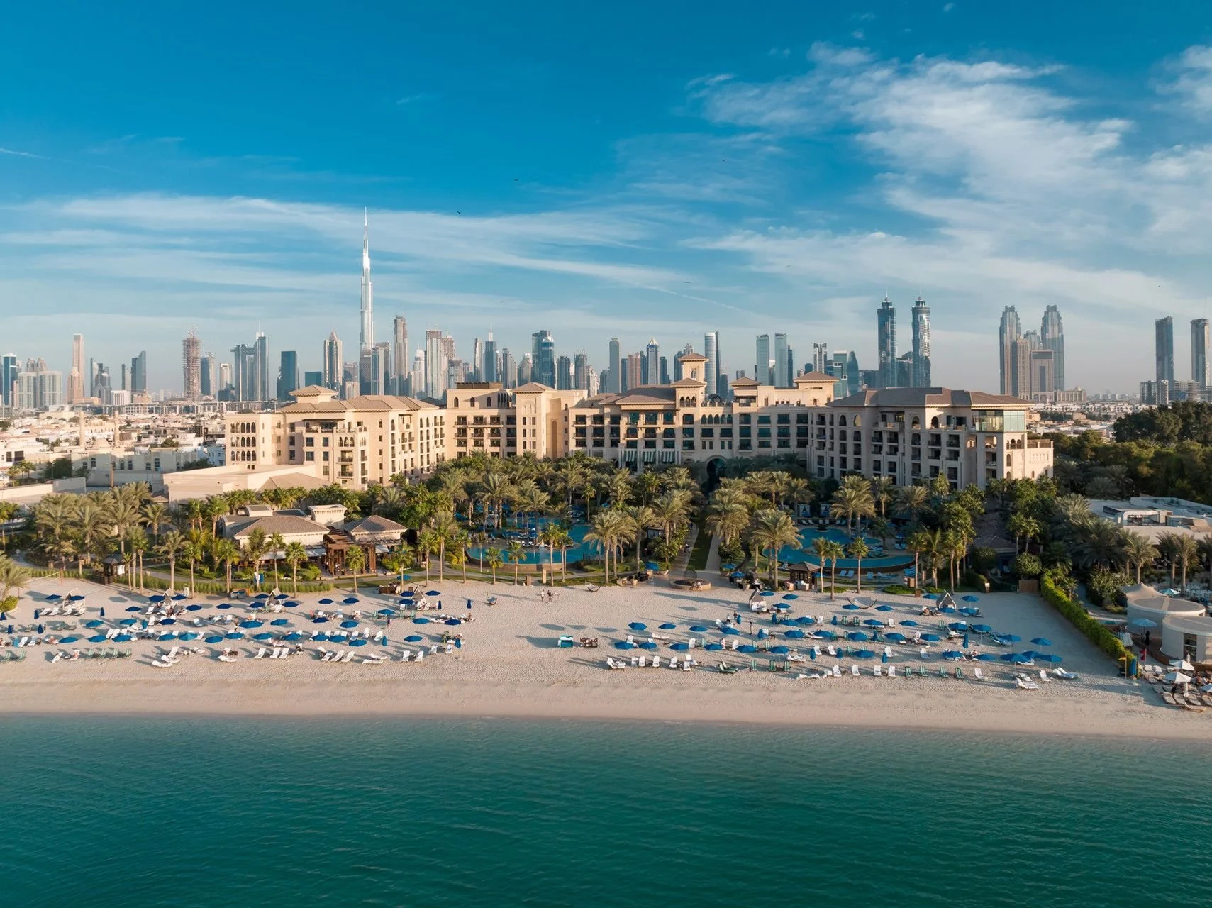 A luxury beach resort with a sandy beach, numerous beach chairs and umbrellas, a swimming pool surrounded by palm trees, and a city skyline with tall skyscrapers including the Burj Khalifa in the background.