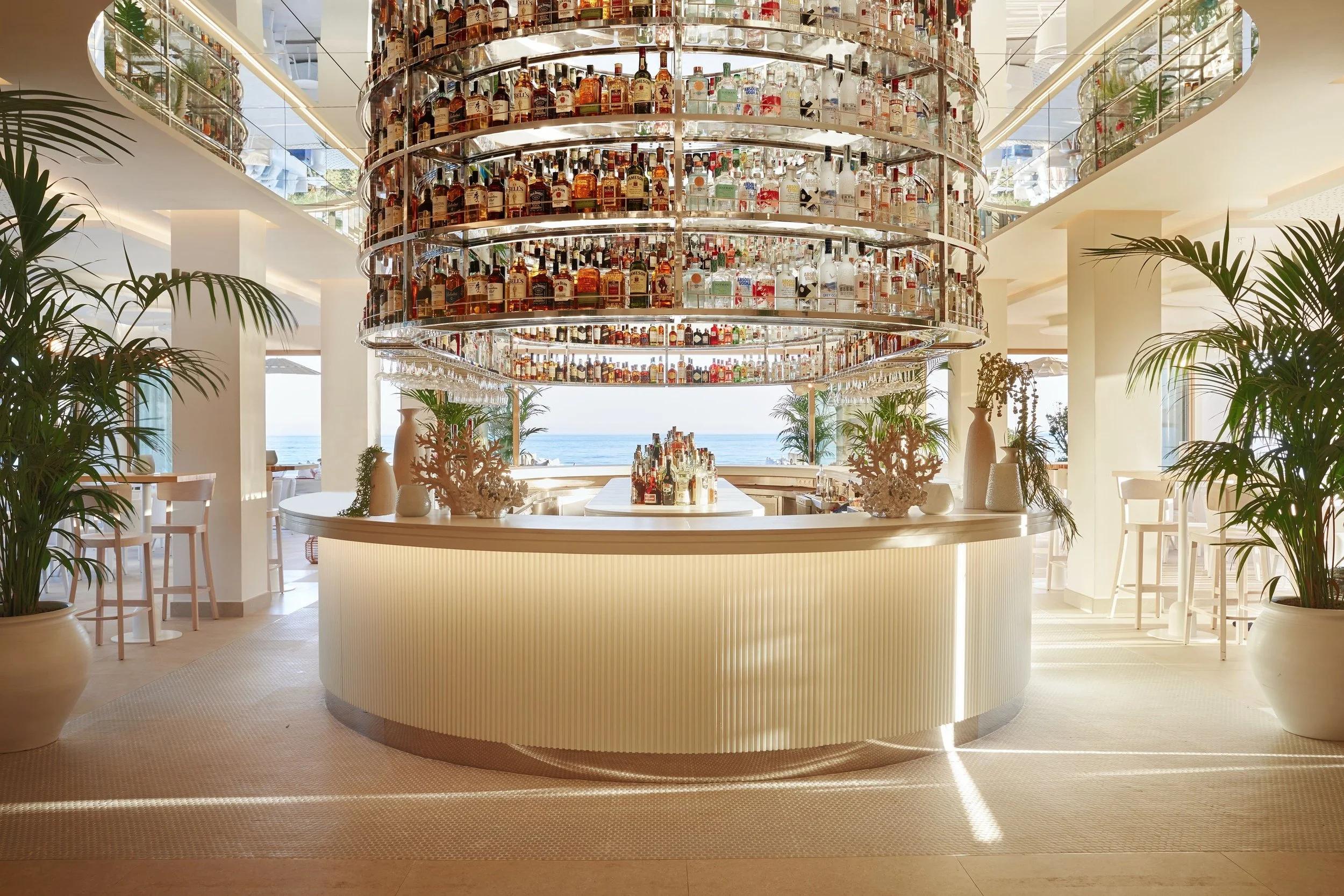 A modern bar area with a large circular bar counter and a tall, cylindrical liquor shelf filled with various bottles of alcohol. The background features large windows overlooking the ocean, and there are potted plants around the space. The decor is light and airy with white walls and furniture.