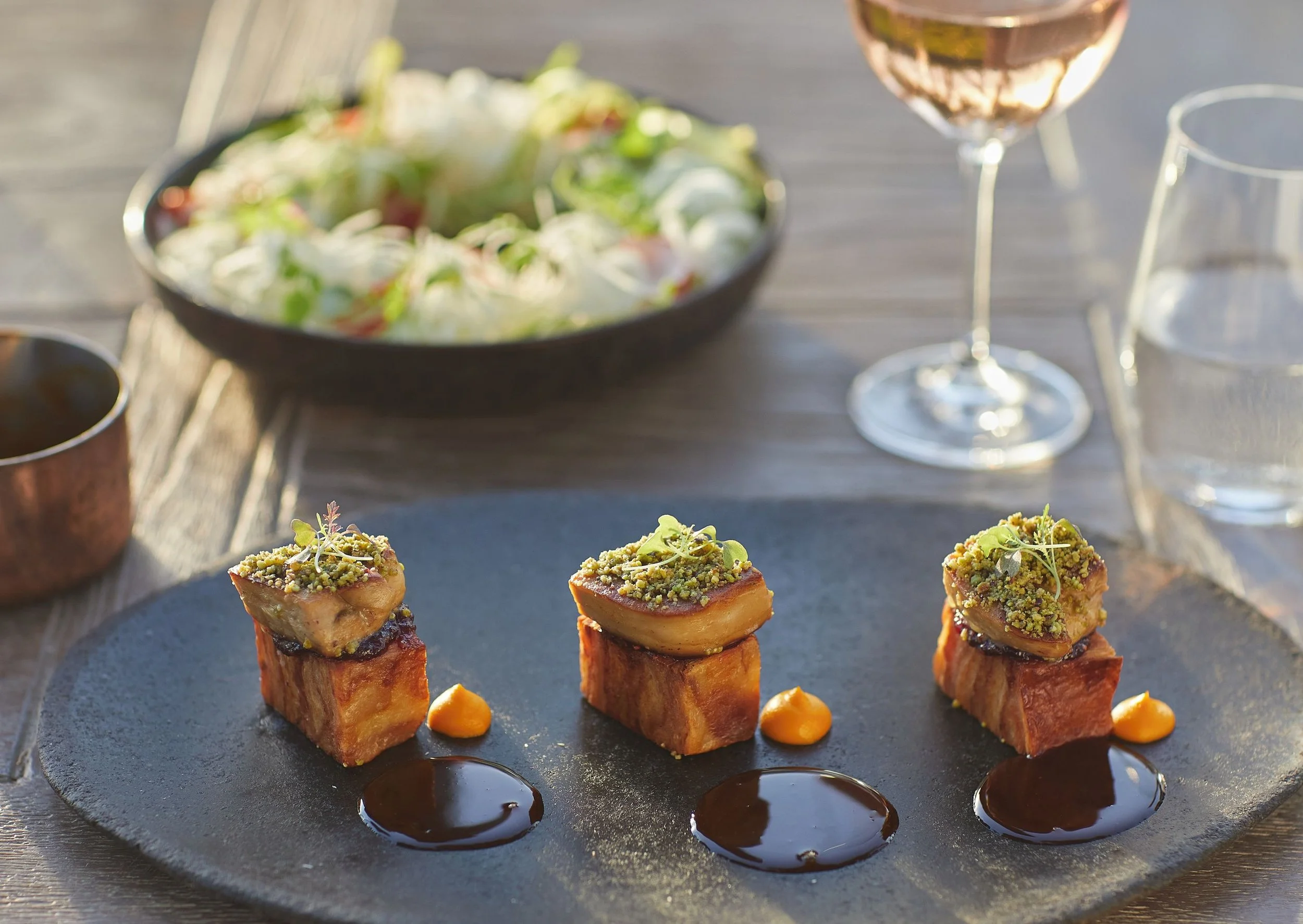 Close-up of four small square food portions topped with chopped herbs and microgreens on a round black slate plate, with dark sauce dots around each portion, and blurred glassware and a bowl in the background.