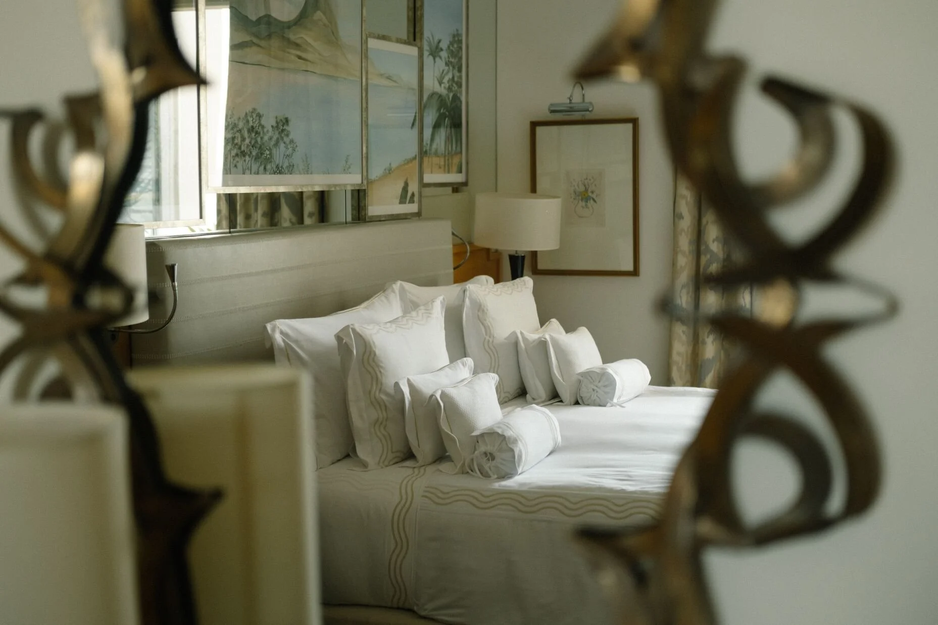 A neatly made bed with white pillows and rolled towels, viewed through an ornate iron gate in a hotel room. There are framed artworks on the walls and a window with patterned curtains.