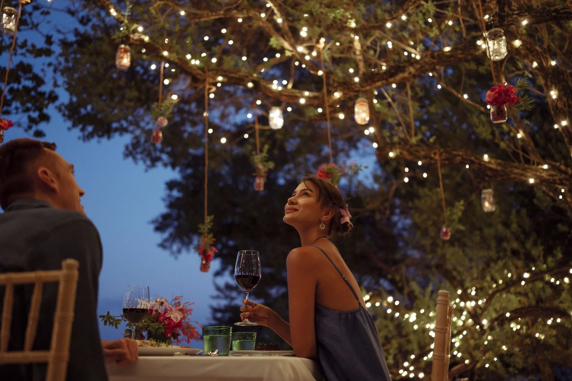 A woman in a blue dress is seated at an outdoor dinner table with a man, holding a glass of red wine. The setting is decorated with hanging jar lanterns and string lights under a large tree. The scene is during evening or nighttime.