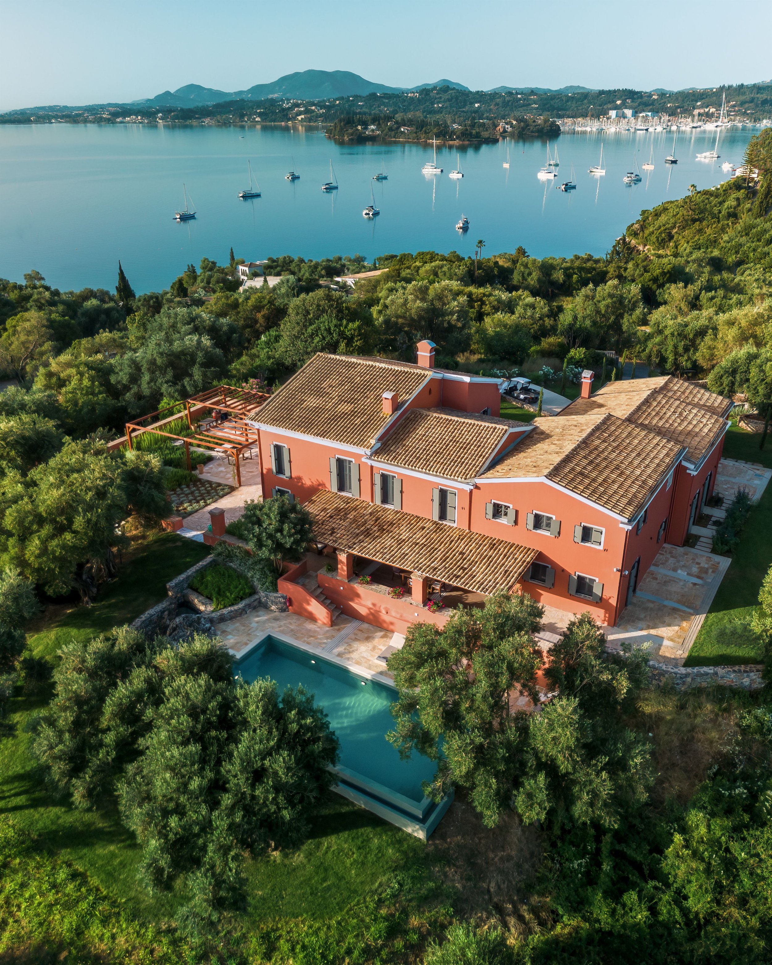Aerial view of a pink house with a terracotta tiled roof surrounded by lush green trees, a swimming pool, and a scenic lake with sailboats in the distance, mountain range in the background.