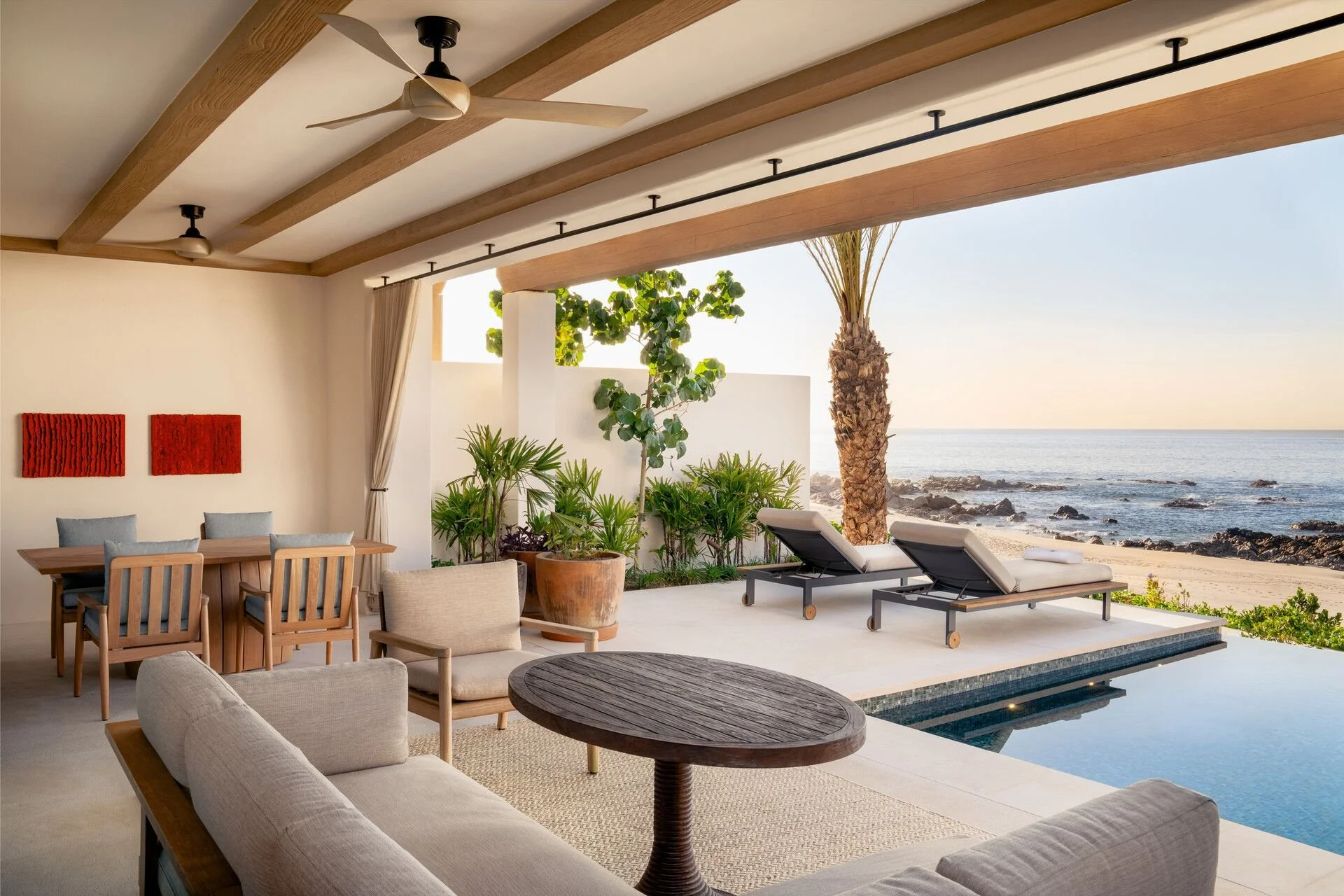 Luxury outdoor living space overlooking the beach with lounge chairs, potted plants, a dining area, and a view of the ocean and rocky shoreline.
