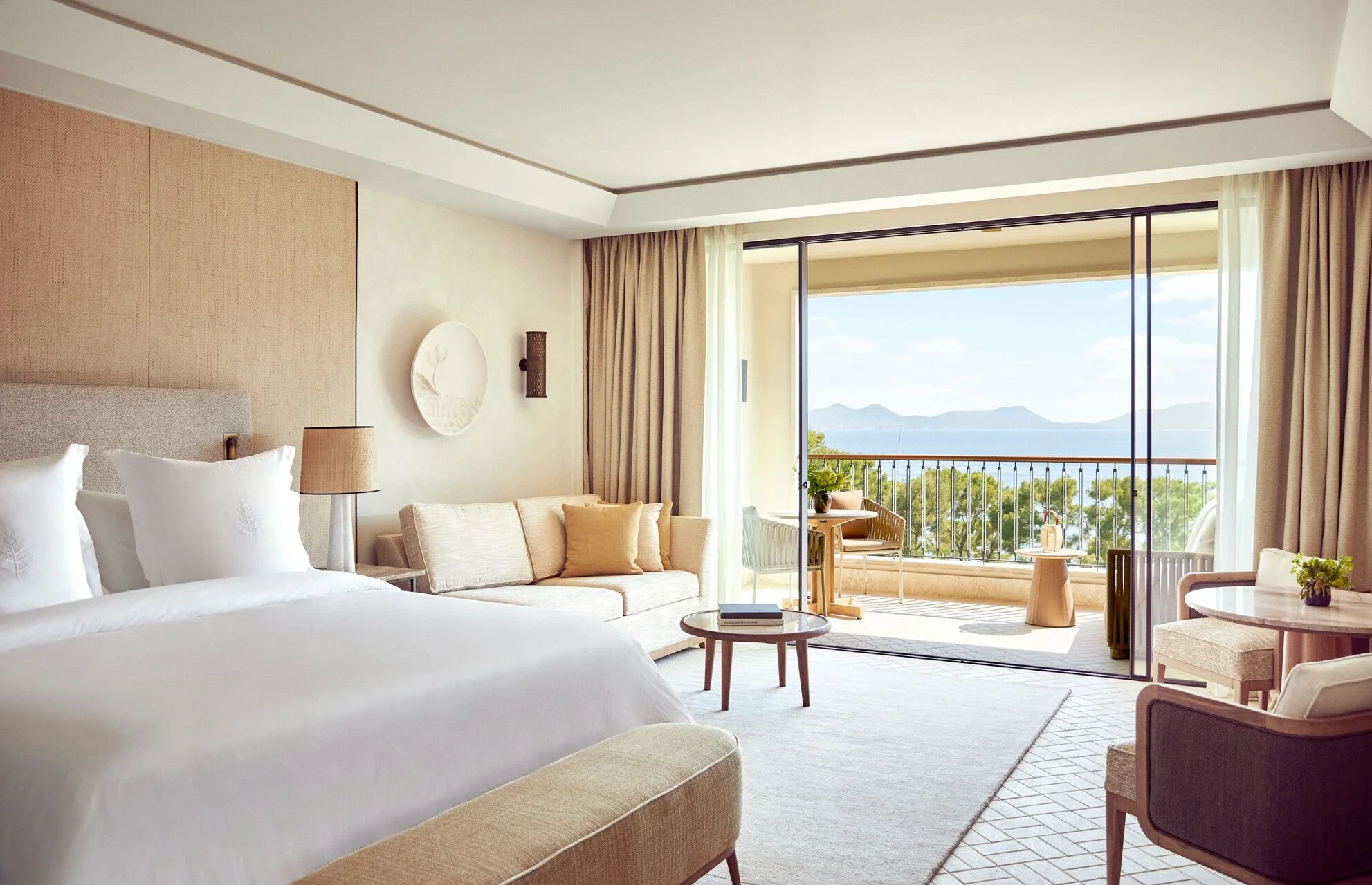 A hotel room with a view of a balcony overlooking water and mountains, featuring a bed, a sofa, a round coffee table, and sliding glass doors with beige curtains.