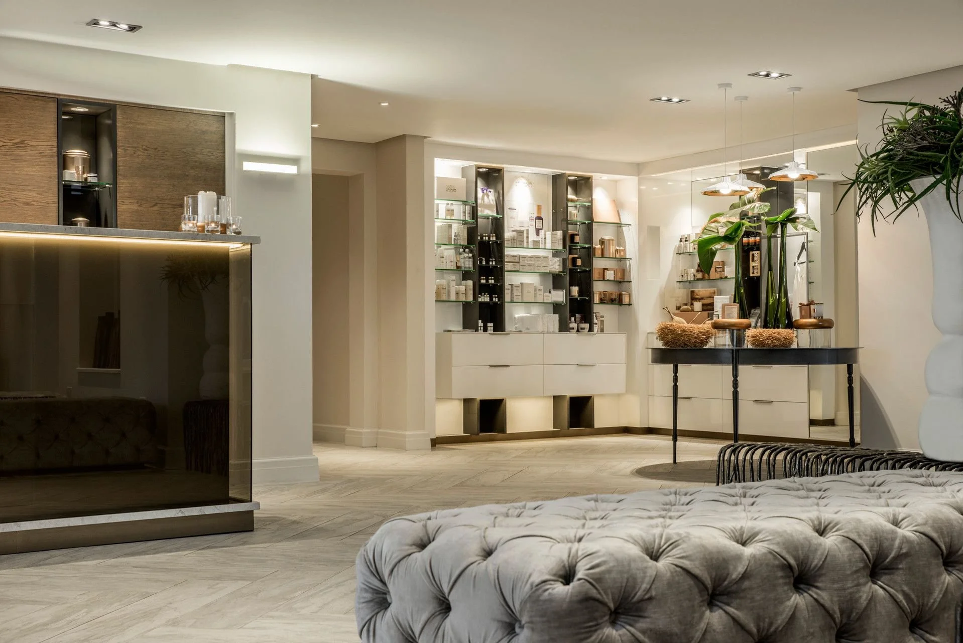 Modern interior space with white walls, wooden and black shelves displaying products, a table with decorative plants, and a tufted beige sofa in the foreground.