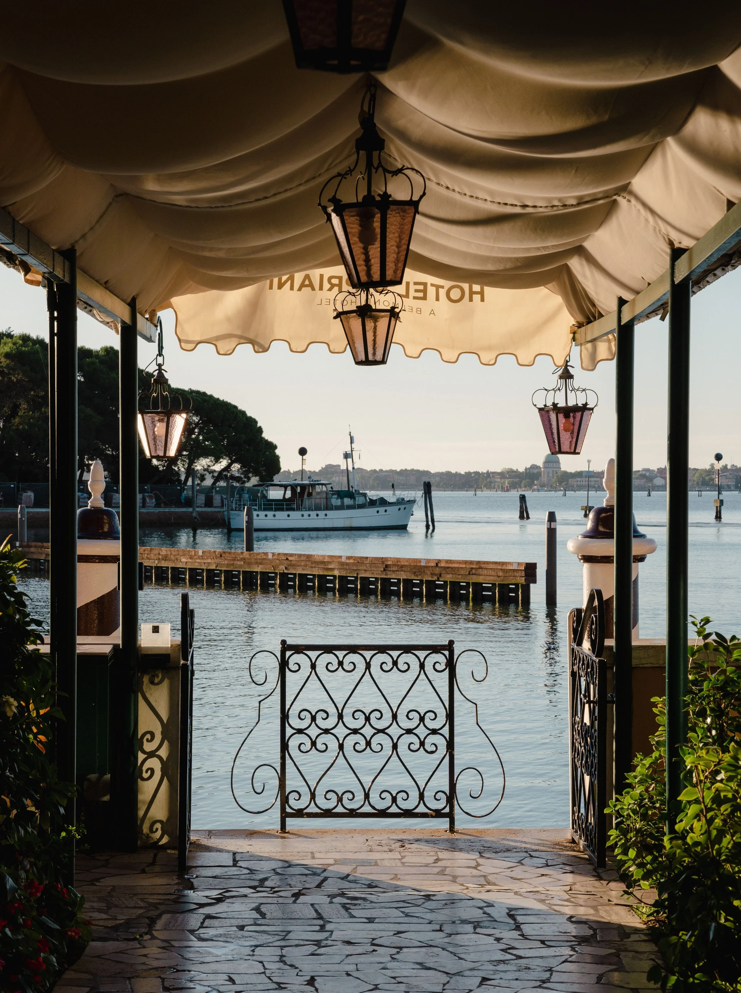 View of a waterfront scene through an ornate wrought-iron gate, with water, a boat, and a city skyline in the distance. A canopy with hanging lanterns shades the entrance.
