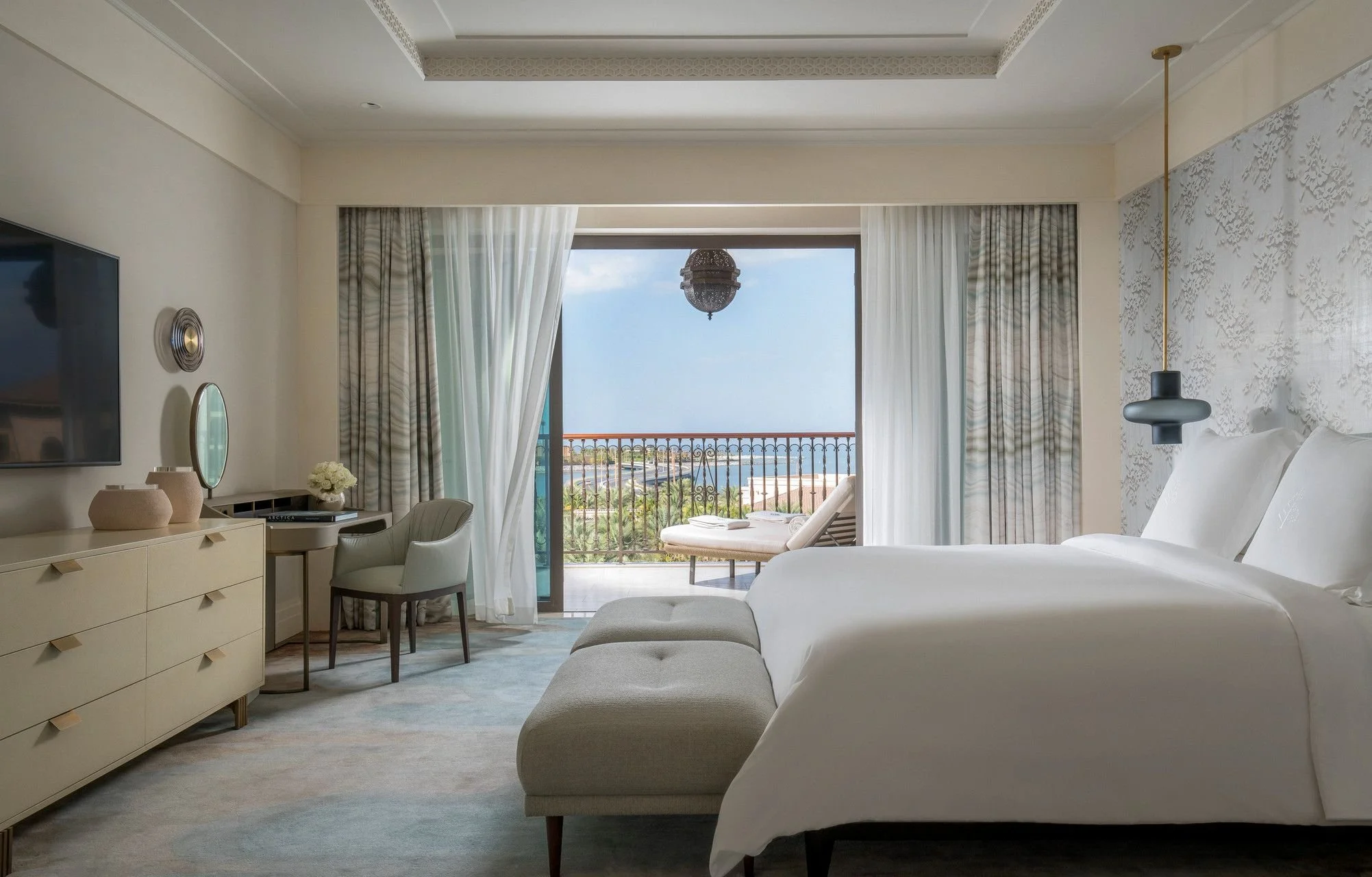 A hotel room with a large bed, a sitting area with chairs and a desk, and a balcony with a view of the ocean and harbor. Curtains are open to reveal the scenic view, and there is a decorative light fixture hanging from the ceiling.