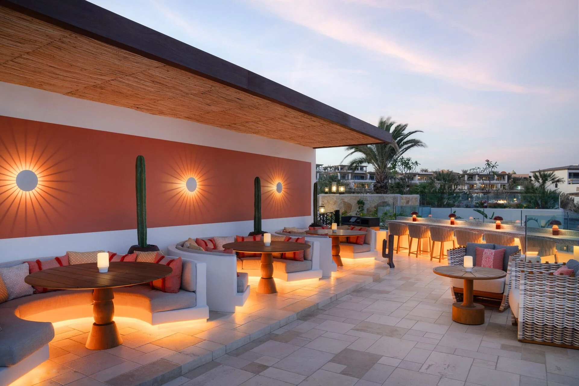 An outdoor lounge area at sunset with modern furniture, wall-mounted lights, and decorative cacti, overlooking a residential neighborhood with palm trees.
