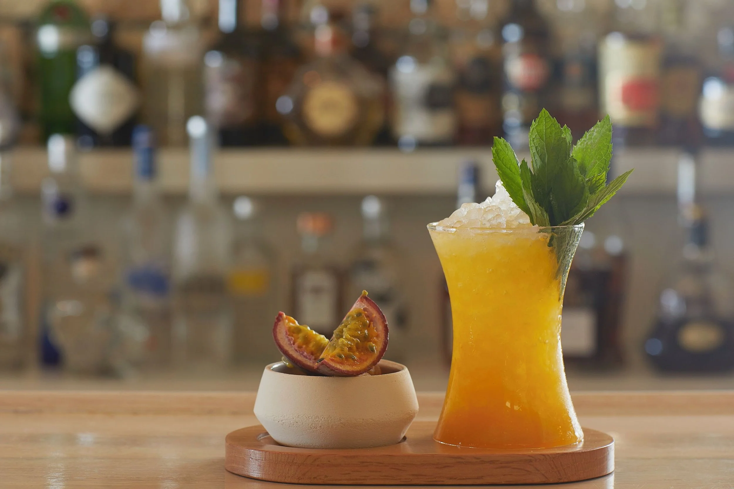 A yellow tropical cocktail garnished with fresh mint leaves in a tall glass, with passion fruit and a small bowl of chopped fruit on a wooden tray, set against a blurred background of bottles.