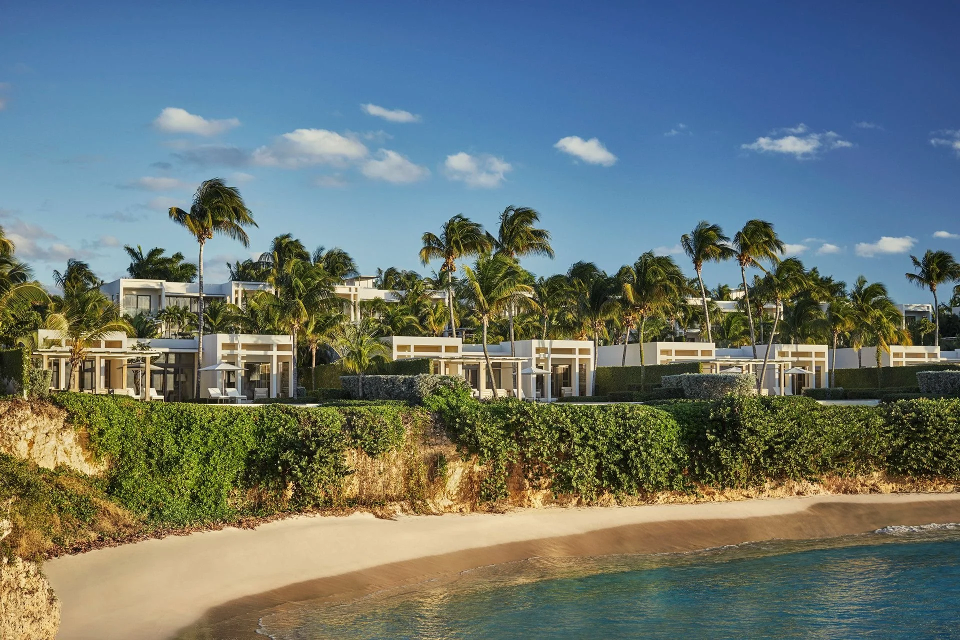 Luxury beachfront resort with modern white buildings, tall palm trees, lush greenery, sandy shoreline, and blue ocean under partly cloudy sky.