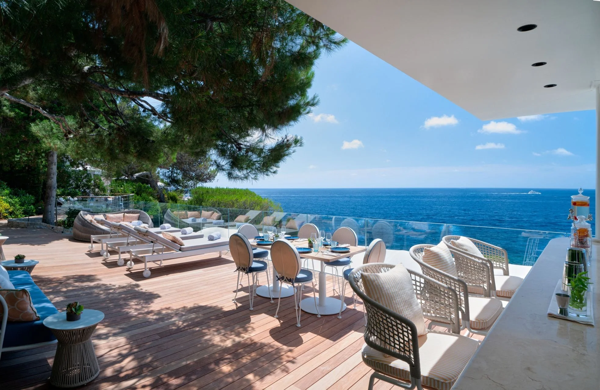 A scenic outdoor dining area overlooking the ocean with modern furniture, trees, and a clear blue sky.