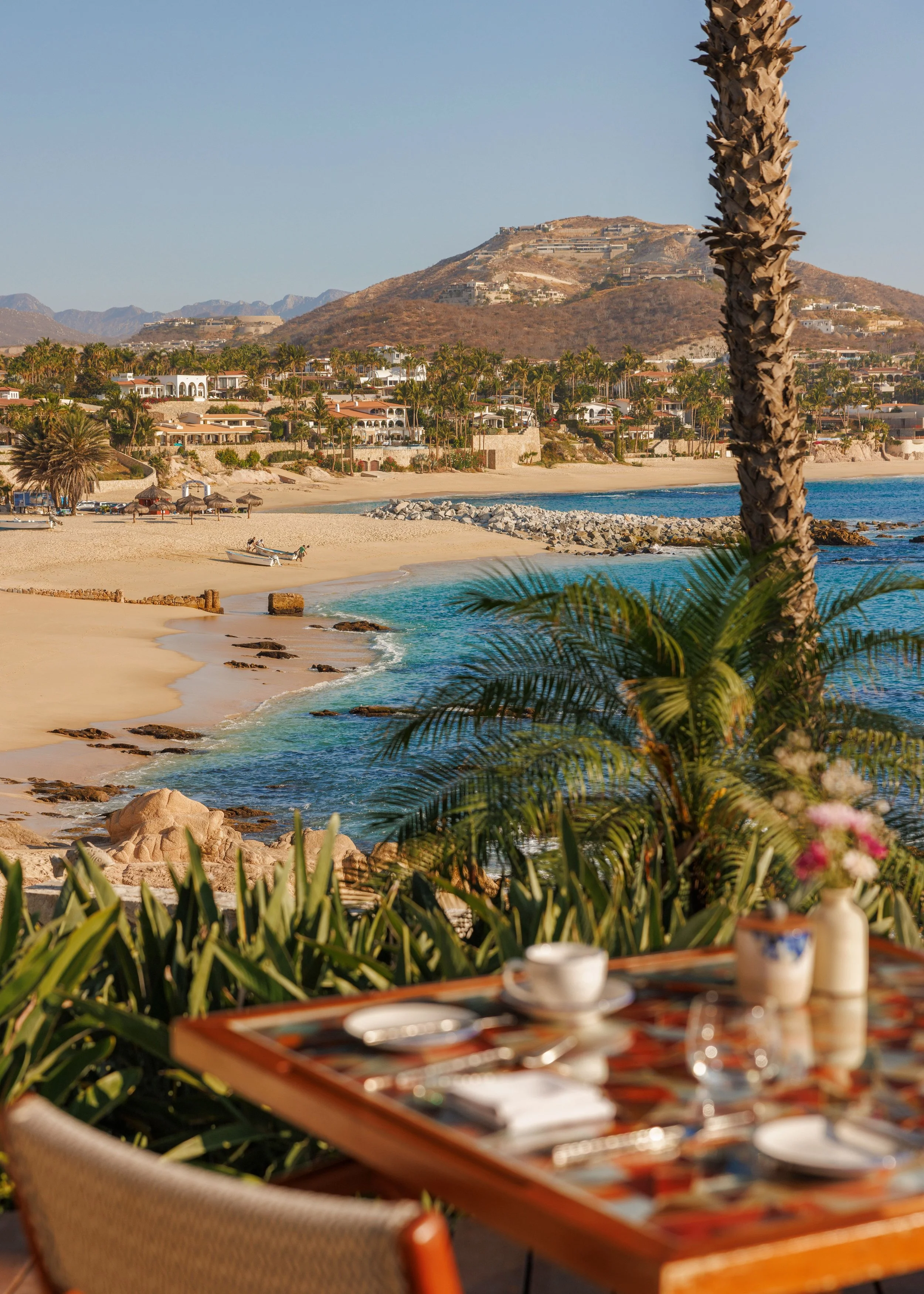 A table set for breakfast with plates, cups, and utensils, overlooking a sandy beach with gentle waves, palm trees, and a view of a hillside with houses.