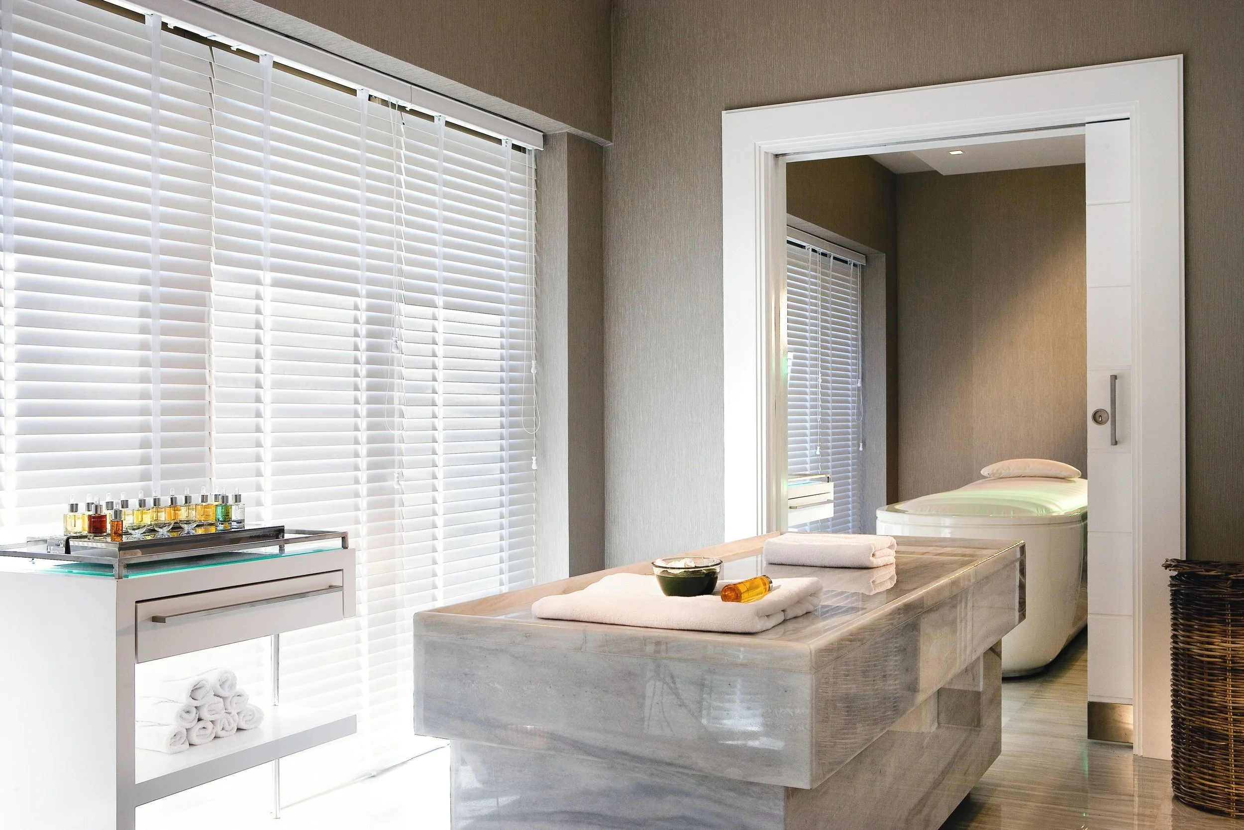 Modern, bright spa or massage room with large window covered by white blinds, marble countertop with neatly folded towels, a bowl, small bottles of essential oil, and a stool. In the background, a doorway reveals a small room with another window and a bed, with neutral-colored walls.