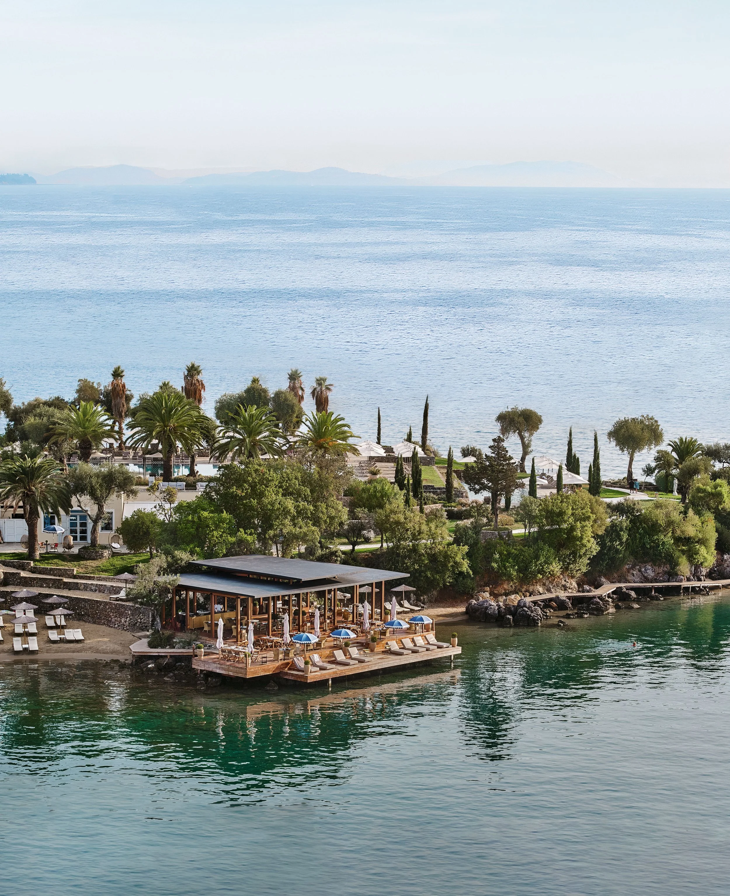 A waterfront scene with palm trees, a terrace with lounge chairs, umbrellas, and a swimming pool, overlooking a large body of water with distant islands and mountains in the background.