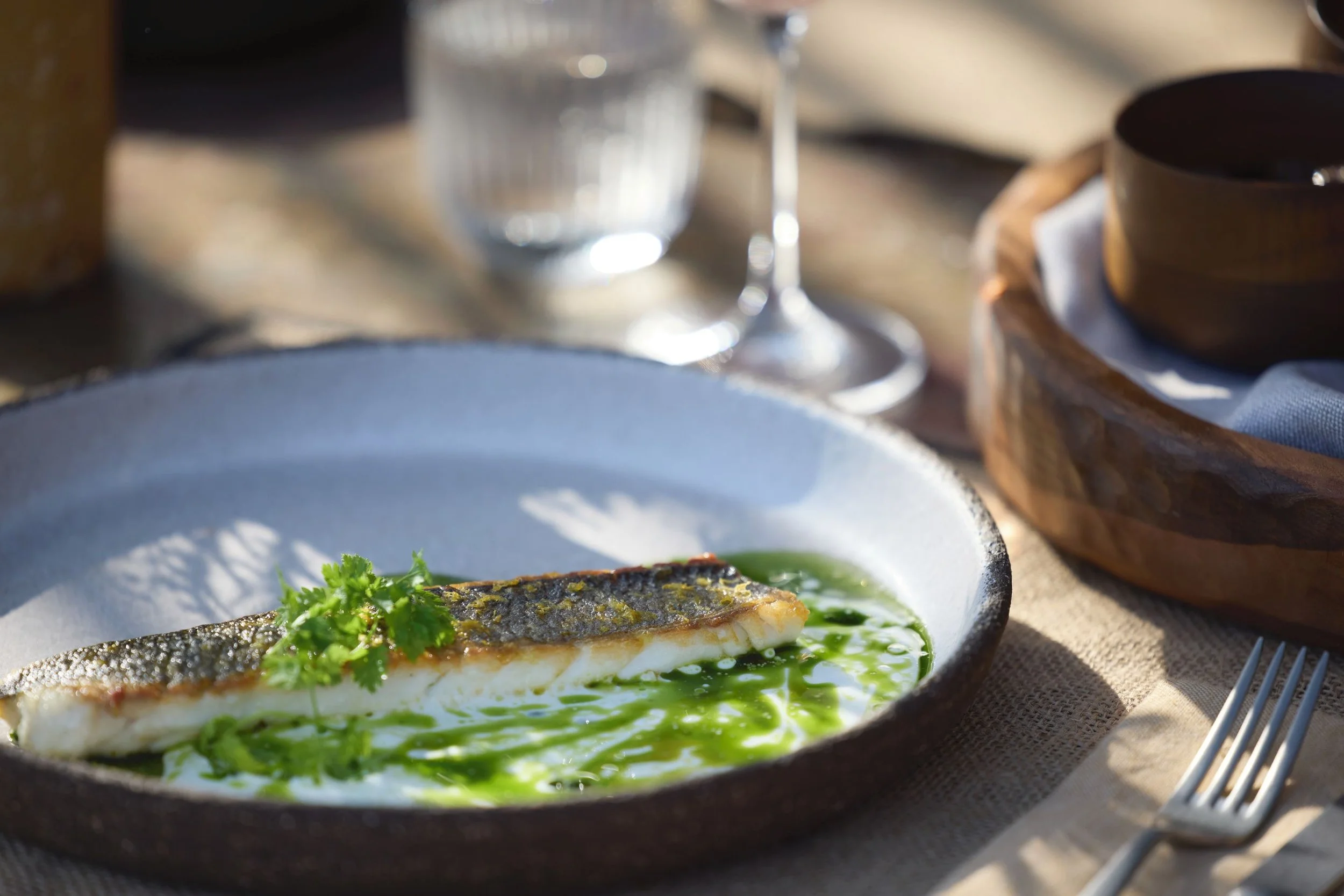 A plated dish featuring a piece of grilled fish garnished with fresh herbs, served with a green sauce, on a dark ceramic plate with a textured surface.