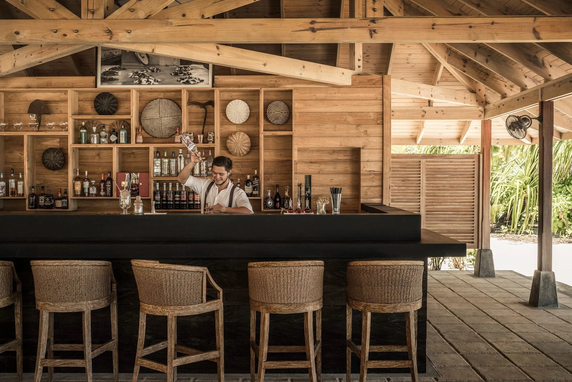 A bartender smiling and pouring alcohol behind a wooden bar, with shelves of bottles and decorative plates on the wood-paneled wall, open-air setting with wicker chairs in front and tropical plants outside.