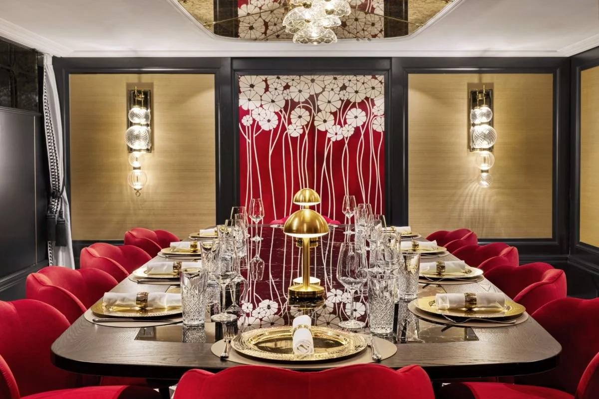 Elegant dining room with a long black table set with gold and glassware, surrounded by red velvet chairs, decorated with gold lamps and modern wall lighting, and a floral wall mural in the background.