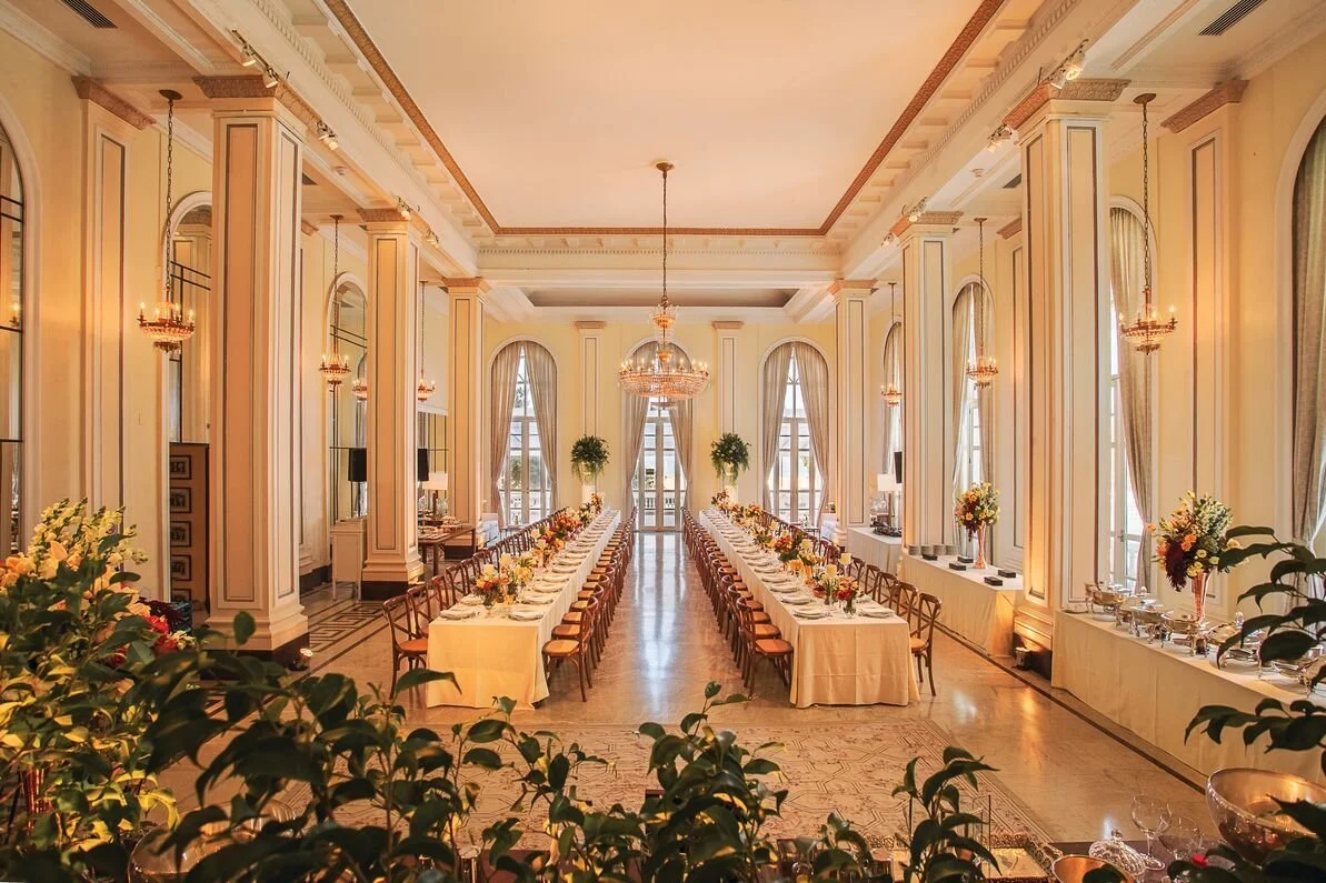 Elegant banquet hall with long tables covered in white tablecloths, decorated with flower arrangements, in a beautifully decorated room with tall arched windows, hanging chandeliers, and golden accents.