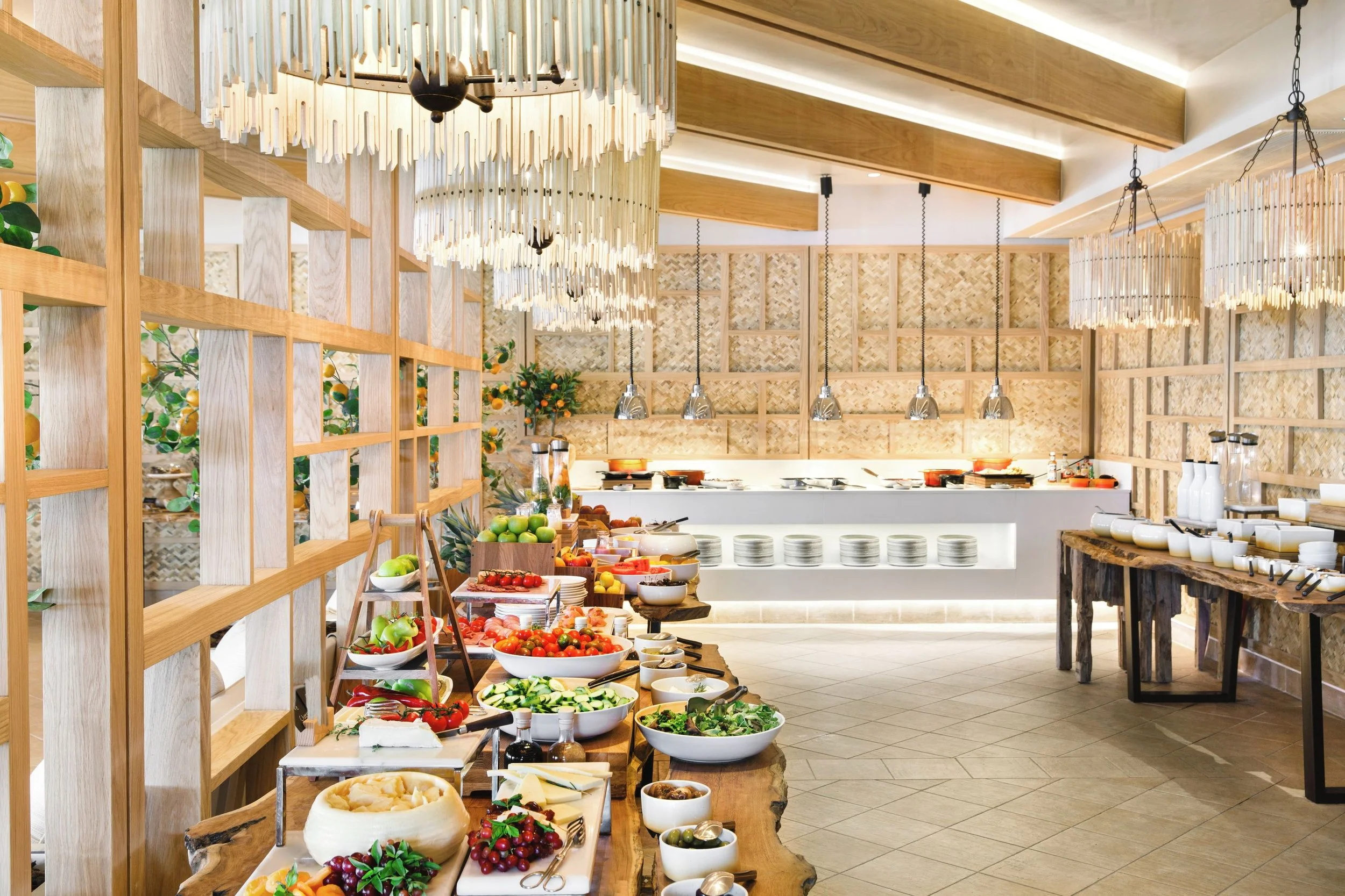 Buffet setup in a modern restaurant with wooden accents, hanging light fixtures, and a variety of fresh fruits, vegetables, and breakfast foods on wooden tables.