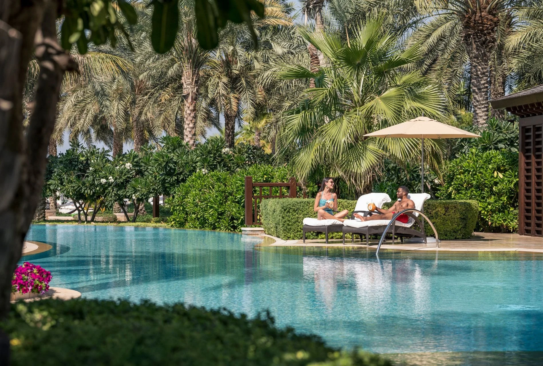 A man and woman relax on lounge chairs by a swimming pool, chatting under a large umbrella, surrounded by palm trees and lush greenery in a tropical setting.
