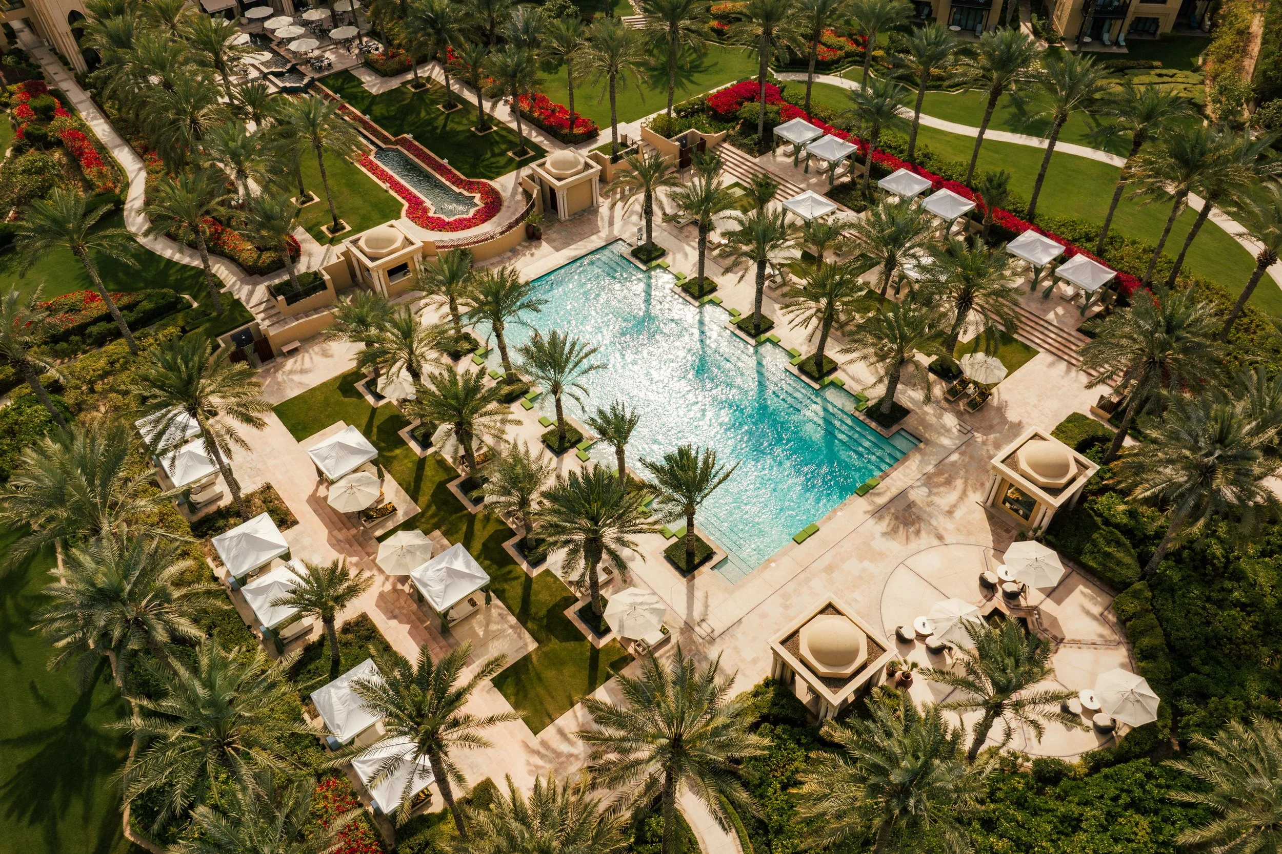 Aerial view of a luxurious hotel pool area with palm trees, white umbrellas, lounging chairs, and well-maintained gardens.