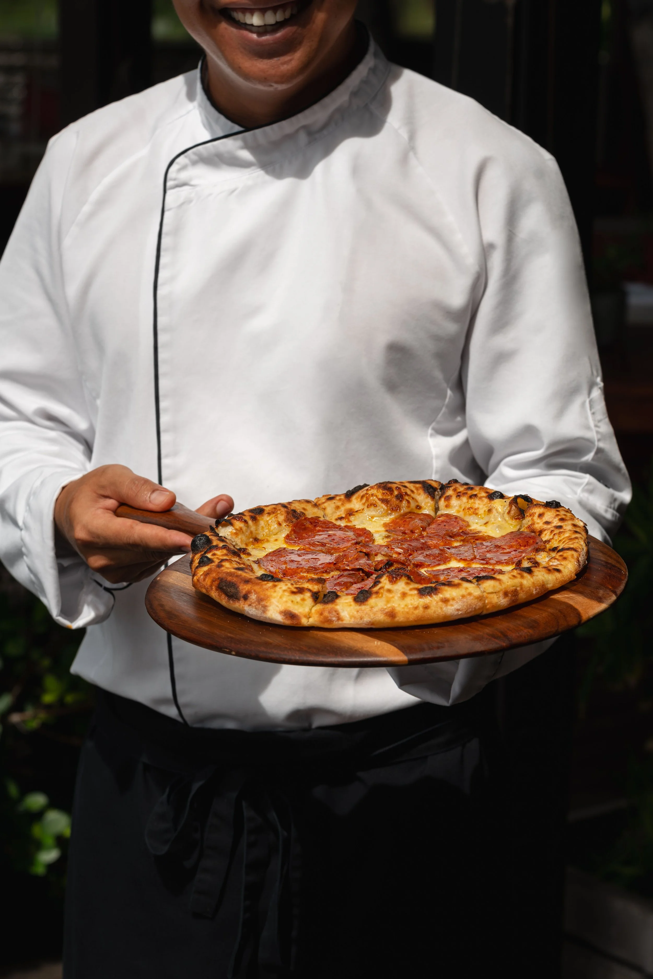 A chef holding a wooden pizza paddle with a freshly baked pepperoni pizza on it, outdoors in a garden setting.