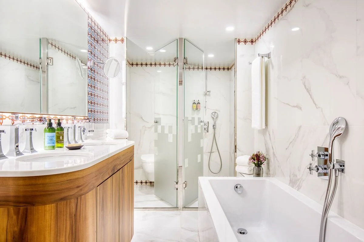 A modern bathroom with a double sink vanity, a glass-enclosed shower, a bathtub, and a toilet, decorated with white marble and colorful tiles.