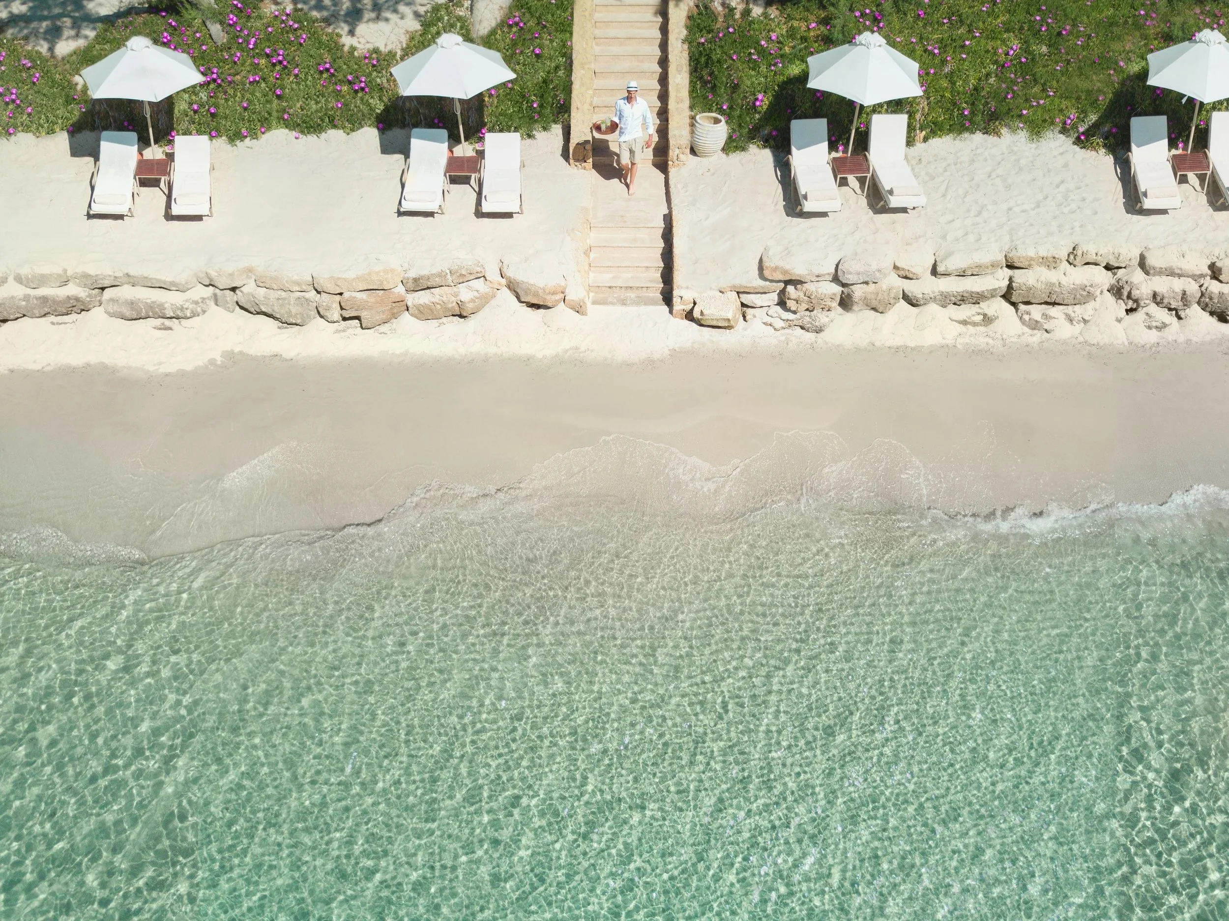 Aerial view of a beachside area with white sand, clear greenish water, and a stone wall separating the beach from a terrace with lounge chairs and white umbrellas. There is a person walking up steps on the terrace, which is decorated with pink flowers and greenery.