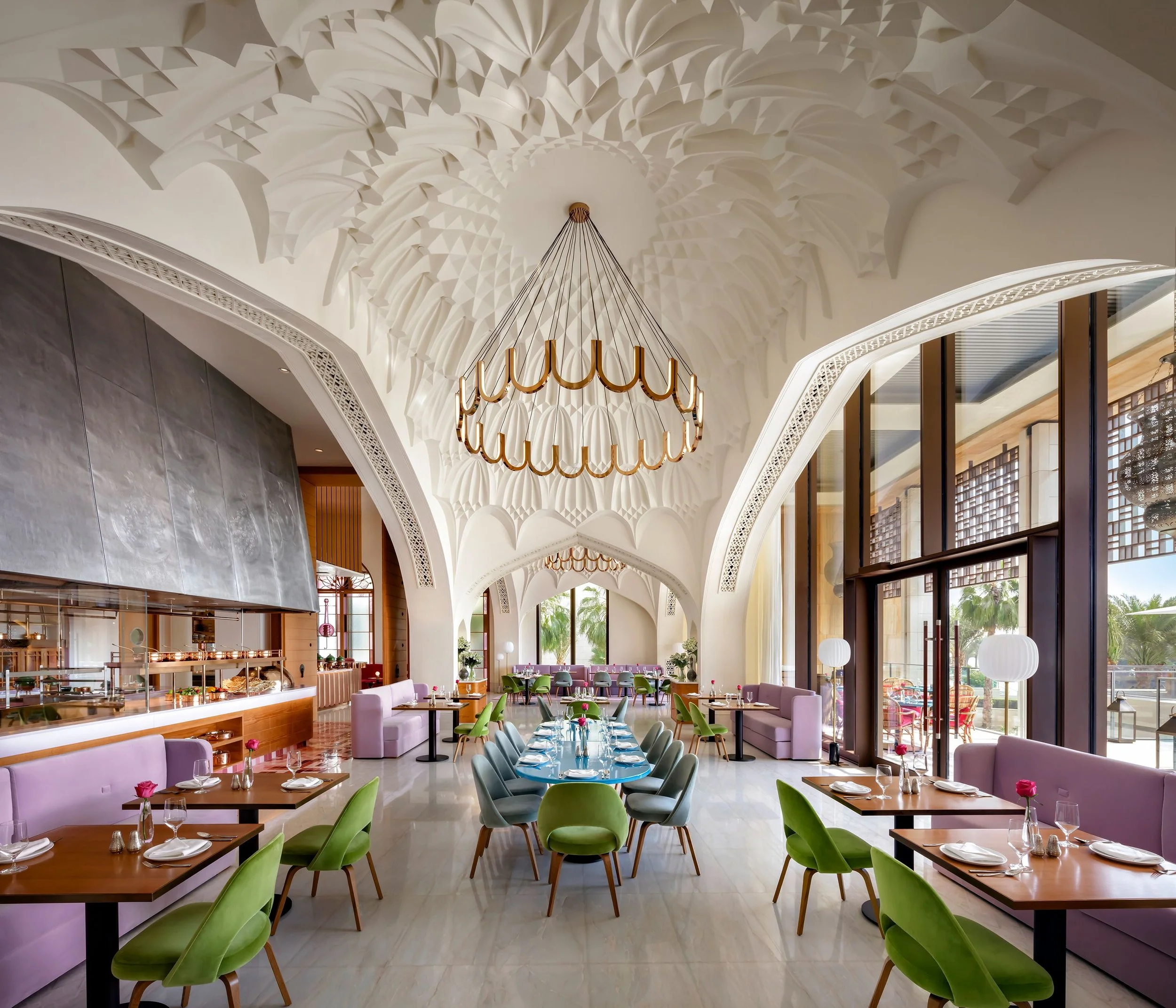 Inside a restaurant with high white decorative ceilings and a large circular chandelier. The dining area has tables set with plates and silverware, surrounded by colorful chairs and soft purple sofas. Large windows let in natural light and outdoor greenery is visible.