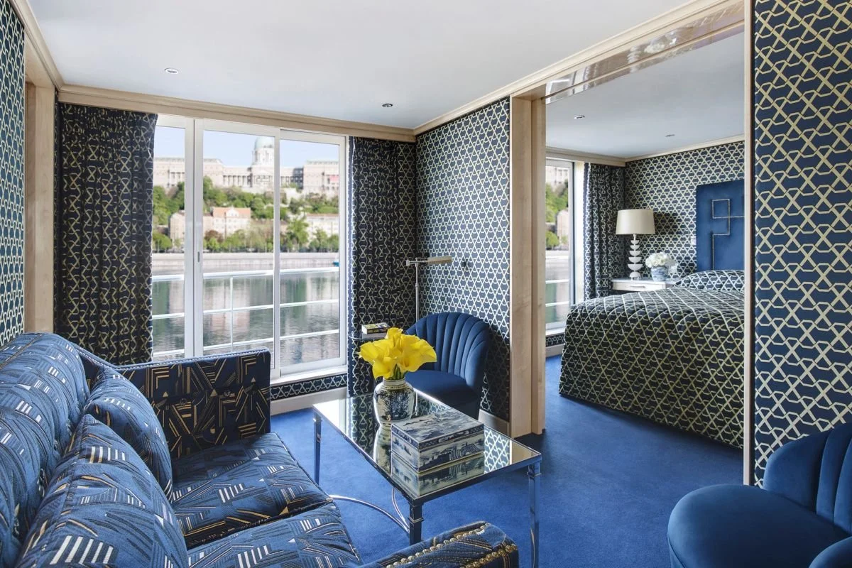 Interior of a hotel suite with blue patterned furniture, a glass coffee table with a vase of yellow flowers, and a bedroom with a matching blue headboard and patterned bedding. Large windows overlook a cityscape across the river.