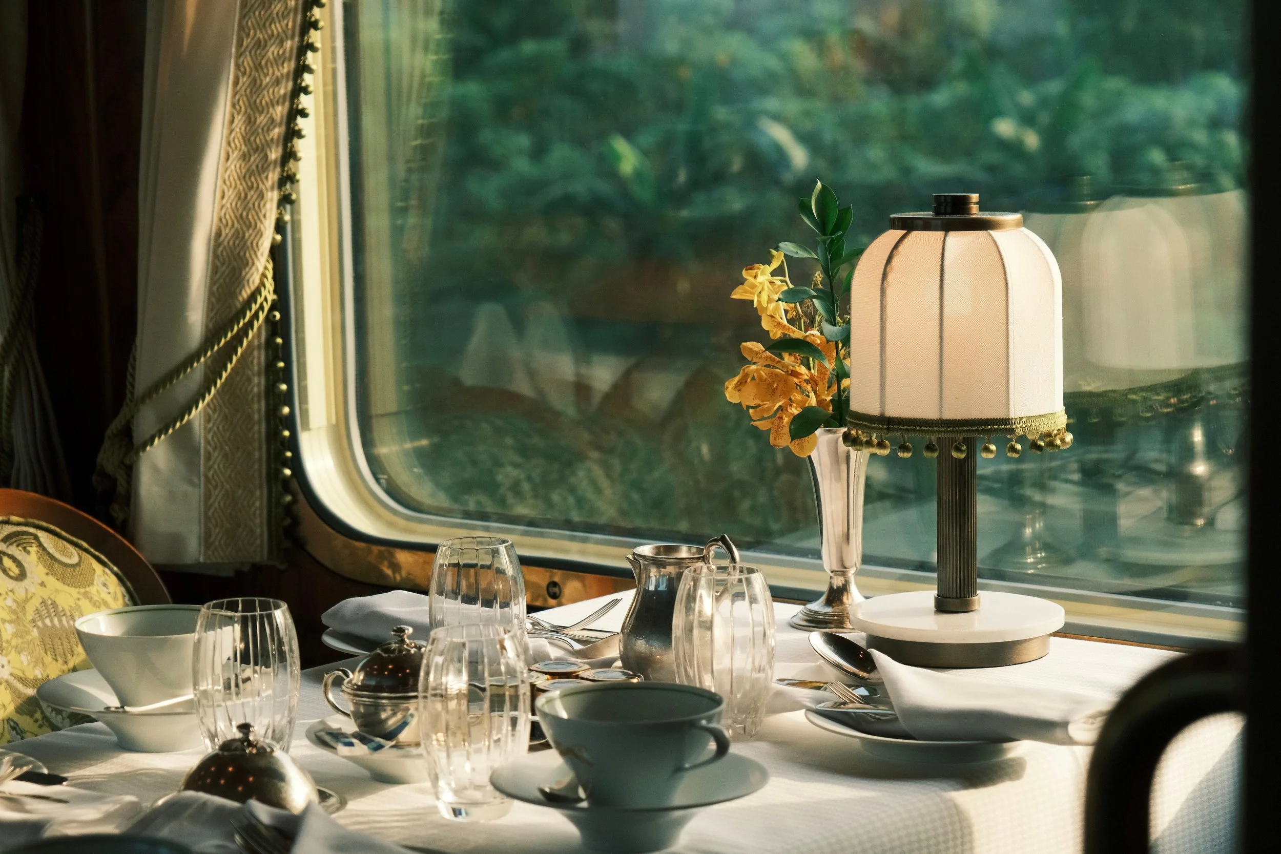 A table set for breakfast inside a train, with various glassware, a bowl, a lamp with a floral decoration, and window view of greenery outside.