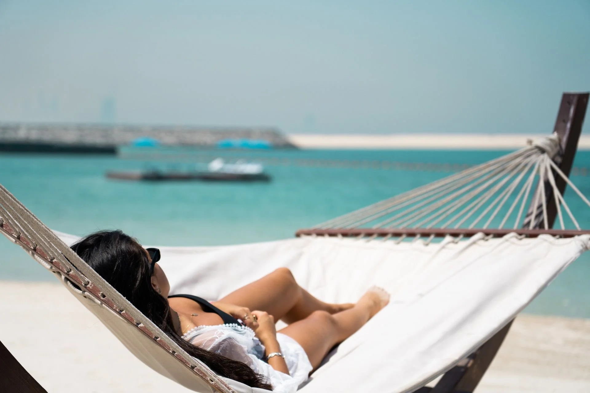 A woman with long dark hair and sunglasses relaxing on a hammock on the beach, with turquoise water and boats in the background.