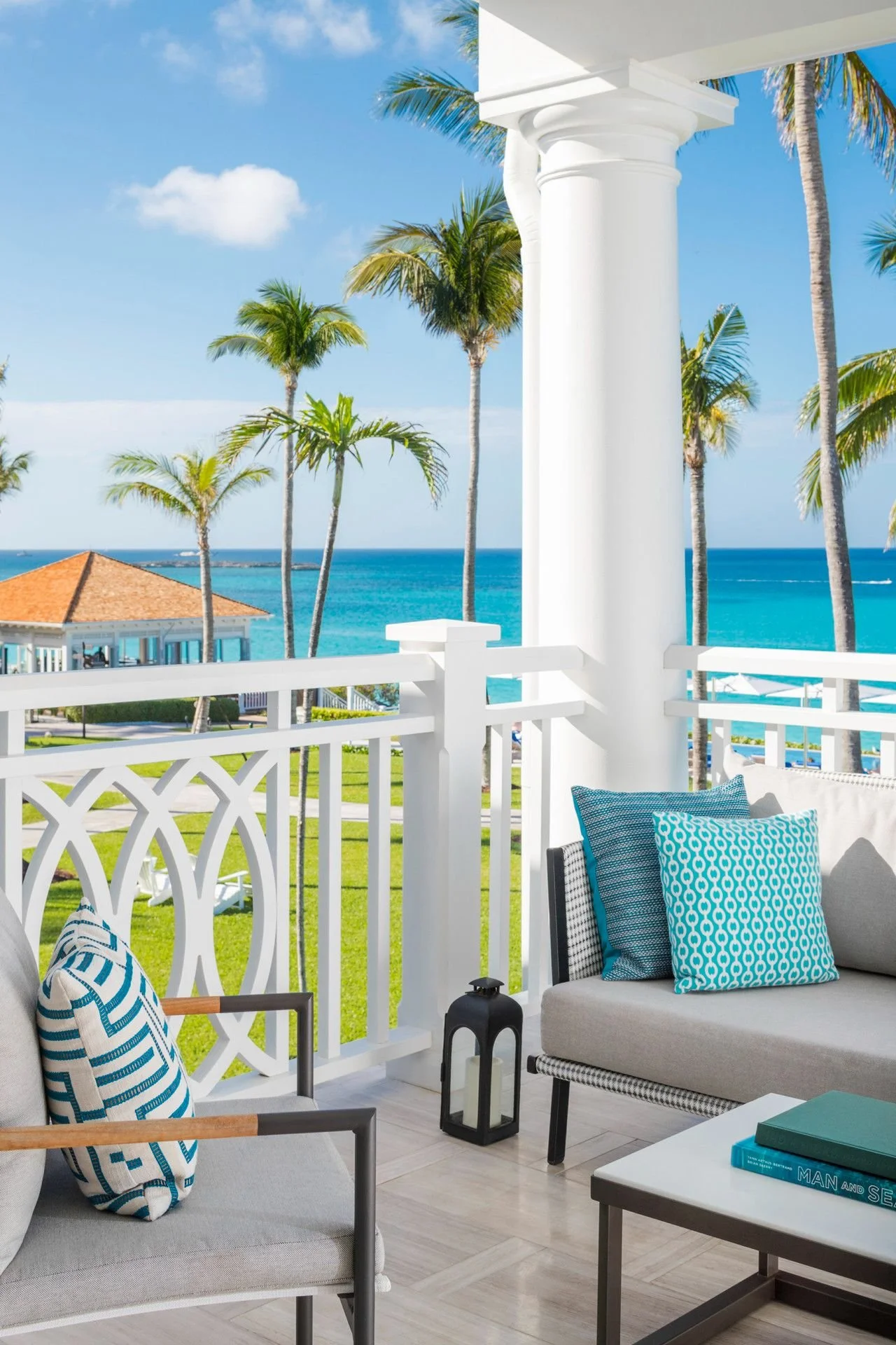Balcony with white railing, tropical view of palm trees, ocean, blue sky with clouds, outdoor seating with blue pillows, green grass