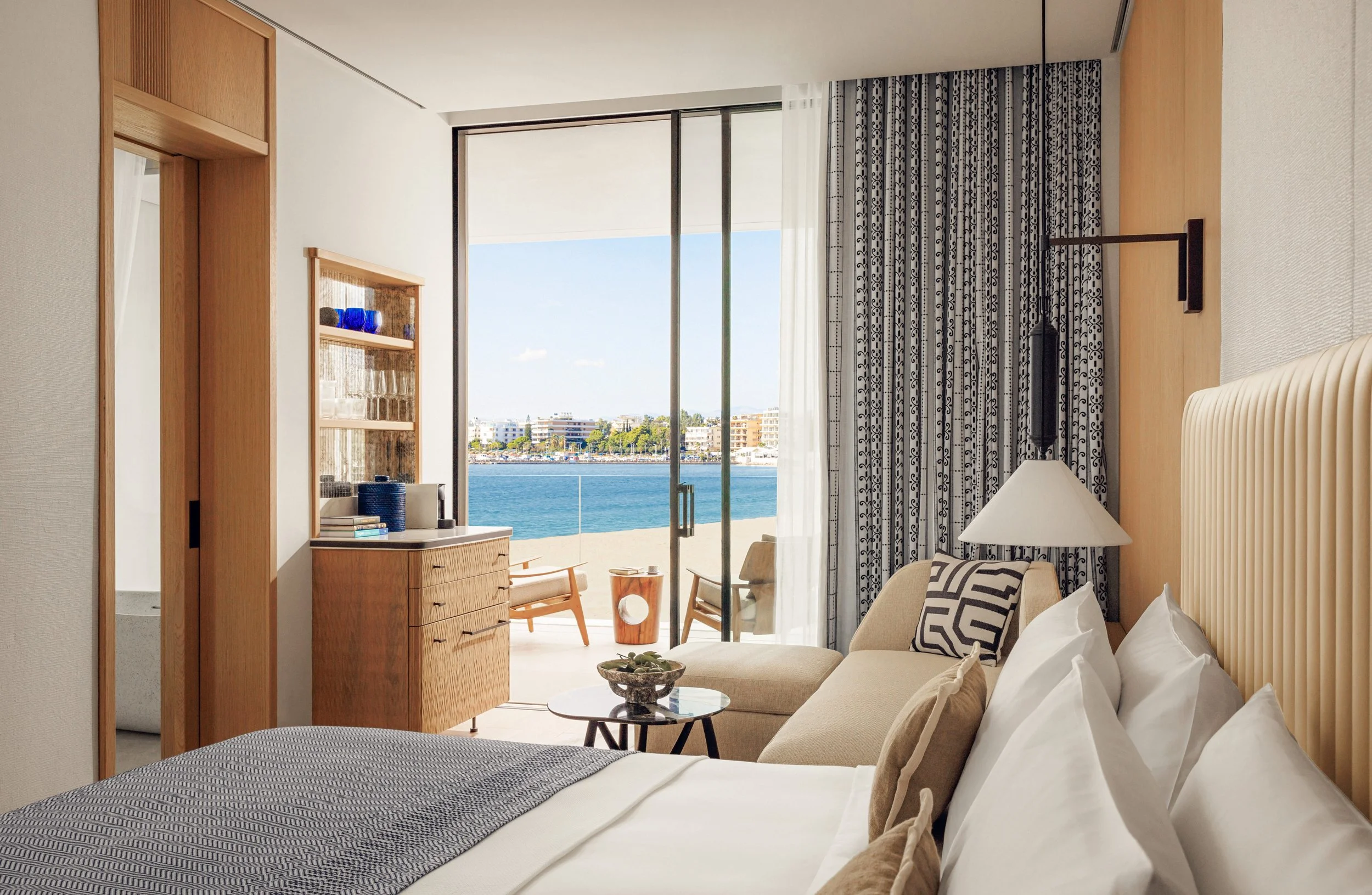Hotel room with a large window showing a balcony and ocean view, decorated in neutral tones with wooden furniture, patterned curtains, and a bed with white bedding.