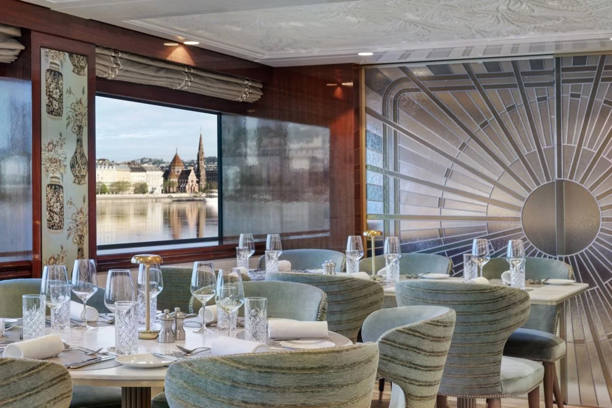 Elegant restaurant with table settings including wine glasses, water glasses, plates, and cutlery, with a large window showing a cityscape and a waterfront.