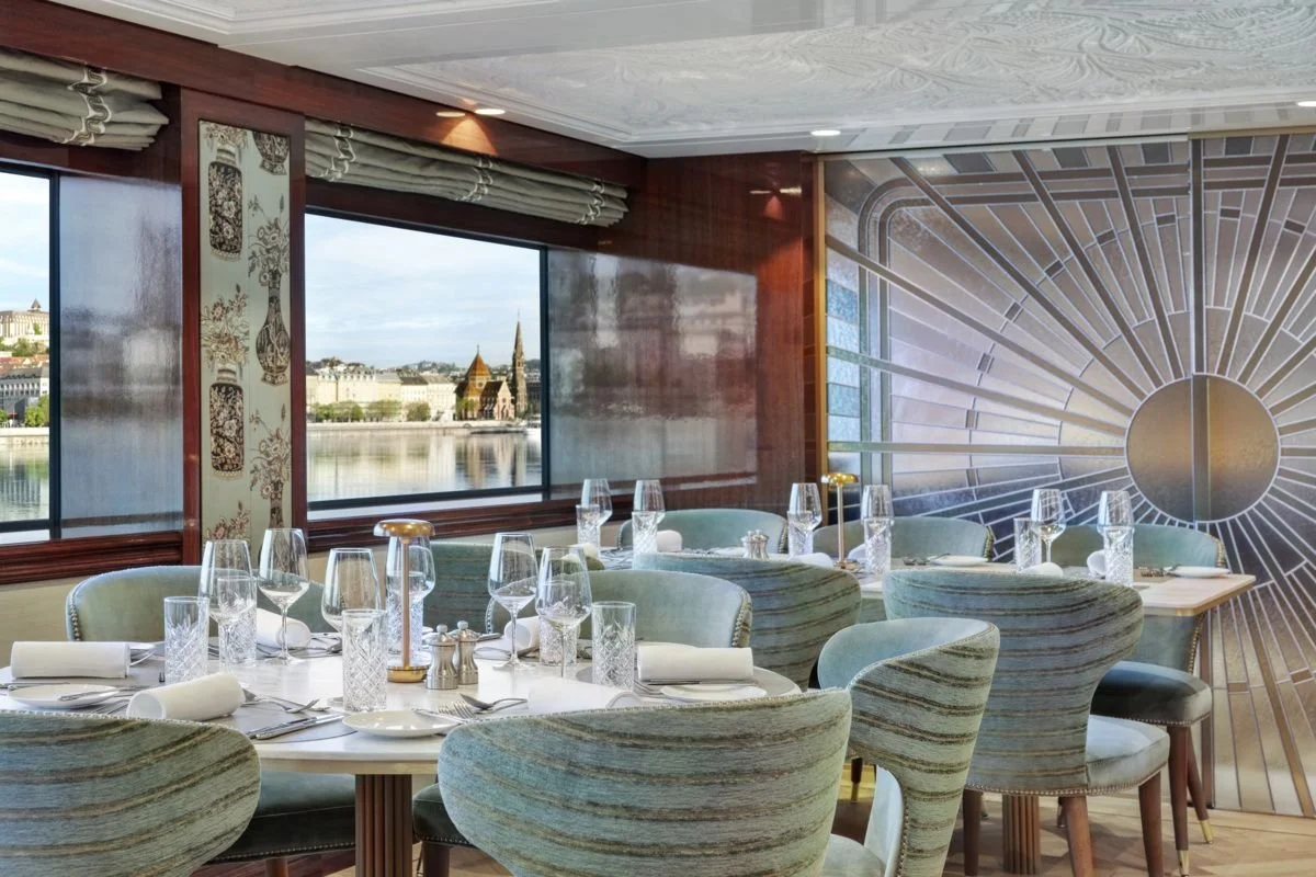 Elegant restaurant dining room with tables set with glassware, silverware, and napkins. Large windows showcase a view of a river and historic buildings across the water. The wall features an artistic geometric sunburst design.