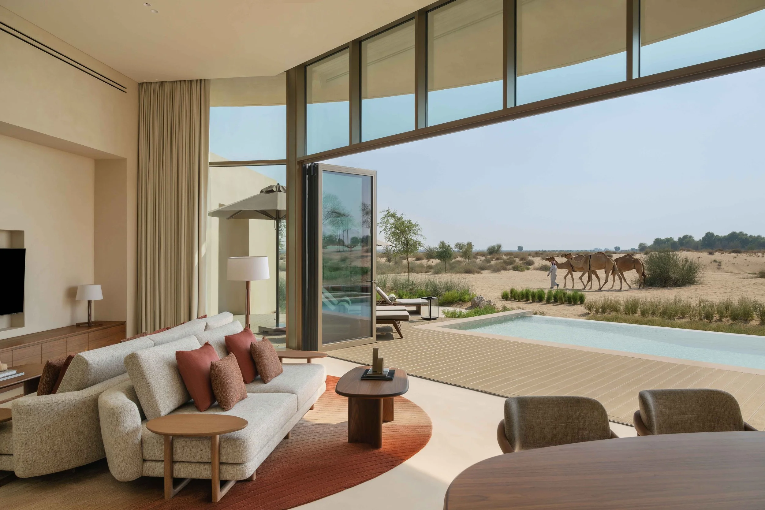 Interior of a modern living room with a large glass wall opening to an outdoor desert landscape. The room features a beige sofa with brown and pink cushions, a wooden coffee table, and a floor lamp. Outside, there are two camels and a person walking in the desert area with sparse vegetation and a clear blue sky.