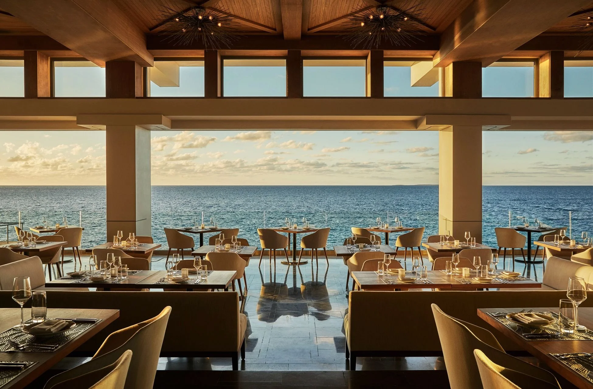 A beachfront restaurant with large windows overlooking the ocean, featuring a wood-paneled ceiling with modern light fixtures, and tables set with glasses and silverware.